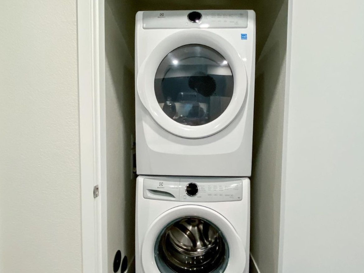 A stacked washer and dryer unit is positioned in a narrow closet space. The washer is located at the bottom, featuring a circular glass door that reveals the drum inside. Above it, the dryer has a similar design with a glass door and control panel at the top. The appliances are white and fit snugly within the closet, with a light-colored wall surrounding them.