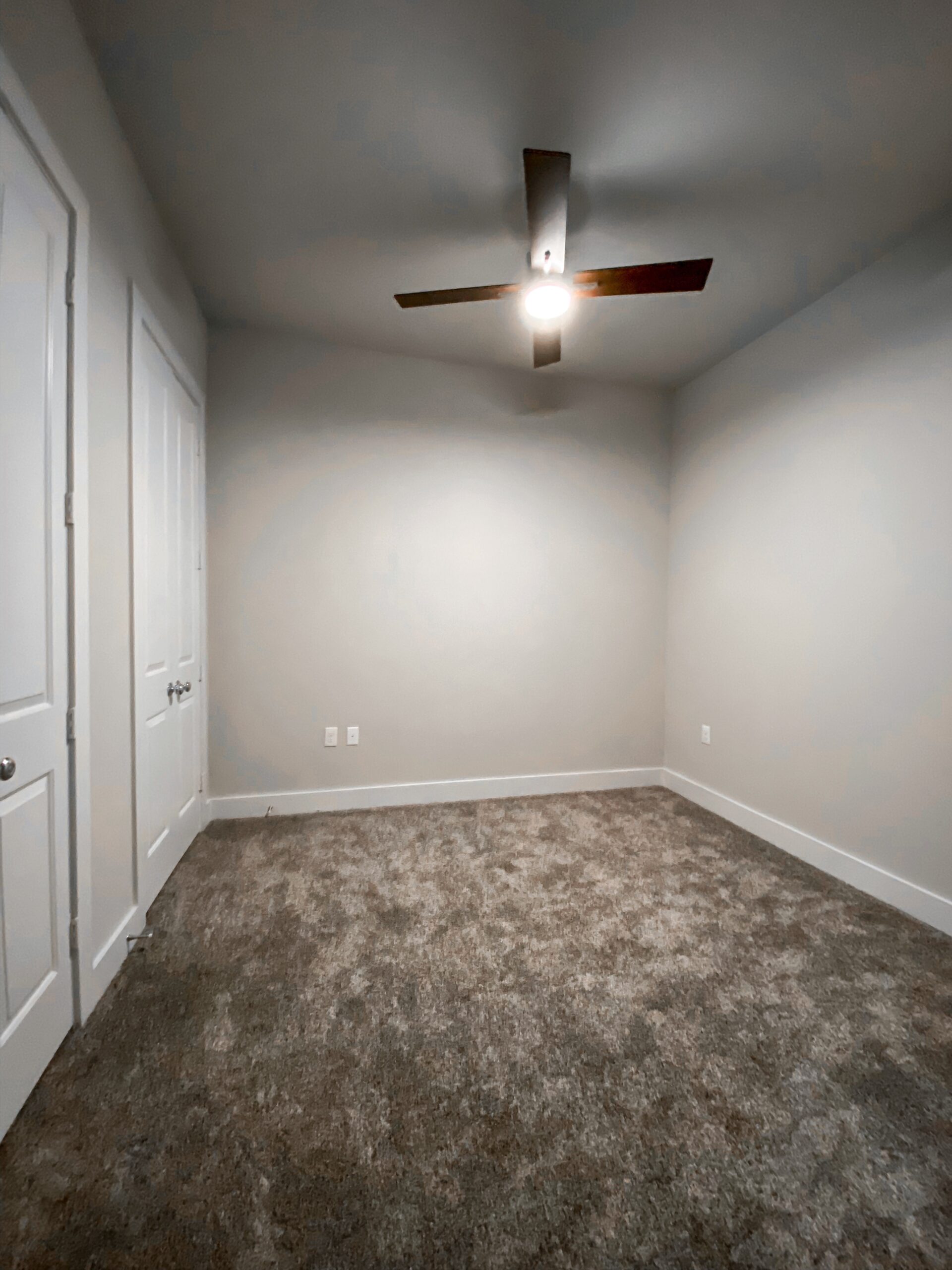 A small, empty room with light gray walls and a plush, beige carpet. On the left side, there are two white doors with silver knobs, likely leading to a closet or storage space. The right wall features two electrical outlets. The overall atmosphere is clean and minimalistic, with ample natural light.