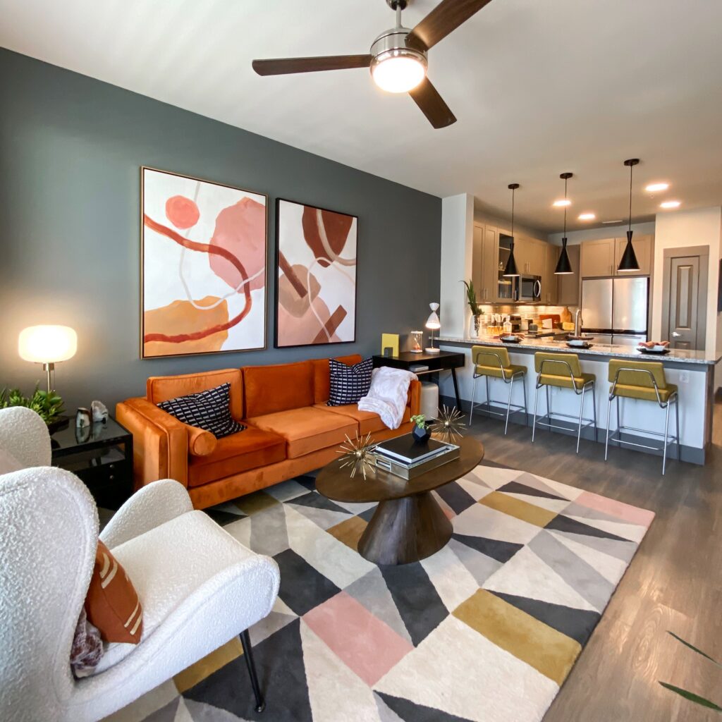 A modern living room features an orange velvet sofa adorned with black and white patterned cushions, accompanied by a cozy white armchair. A round wooden coffee table sits on a geometric area rug with various colors, including pink, gray, and yellow. Two abstract art pieces hang on the dark gray wall above the sofa. In the background, a kitchen area is visible, equipped with stainless steel appliances and bar stools in yellow and chrome. A ceiling fan provides a stylish touch to the space.