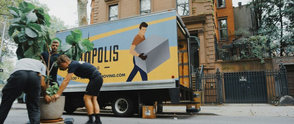A moving truck is parked on a street, featuring a colorful advertisement on its side depicting a person carrying a large box. Two individuals are seen in the foreground, one lifting a potted plant while the other assists. The background includes brownstone buildings with greenery and a "No Parking" sign visible. The scene conveys a busy moving day atmosphere.