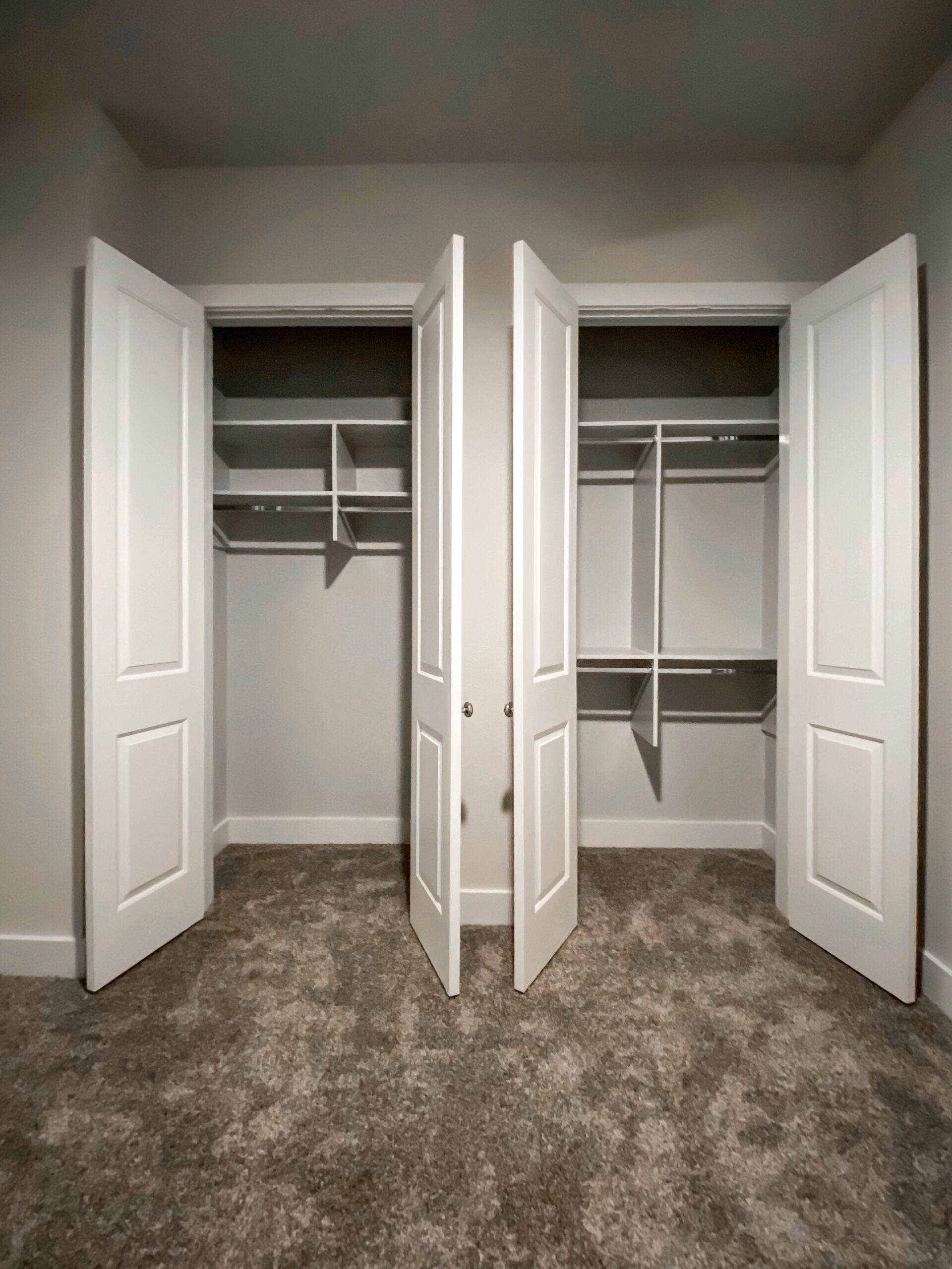 A spacious closet features two sets of double doors, each with a simple, modern design. Inside, the left side has several shelves and a hanging rod, while the right side includes additional shelves and a hanging space. The walls are painted a light color, and the floor is carpeted in a neutral tone, creating a clean and organized appearance.