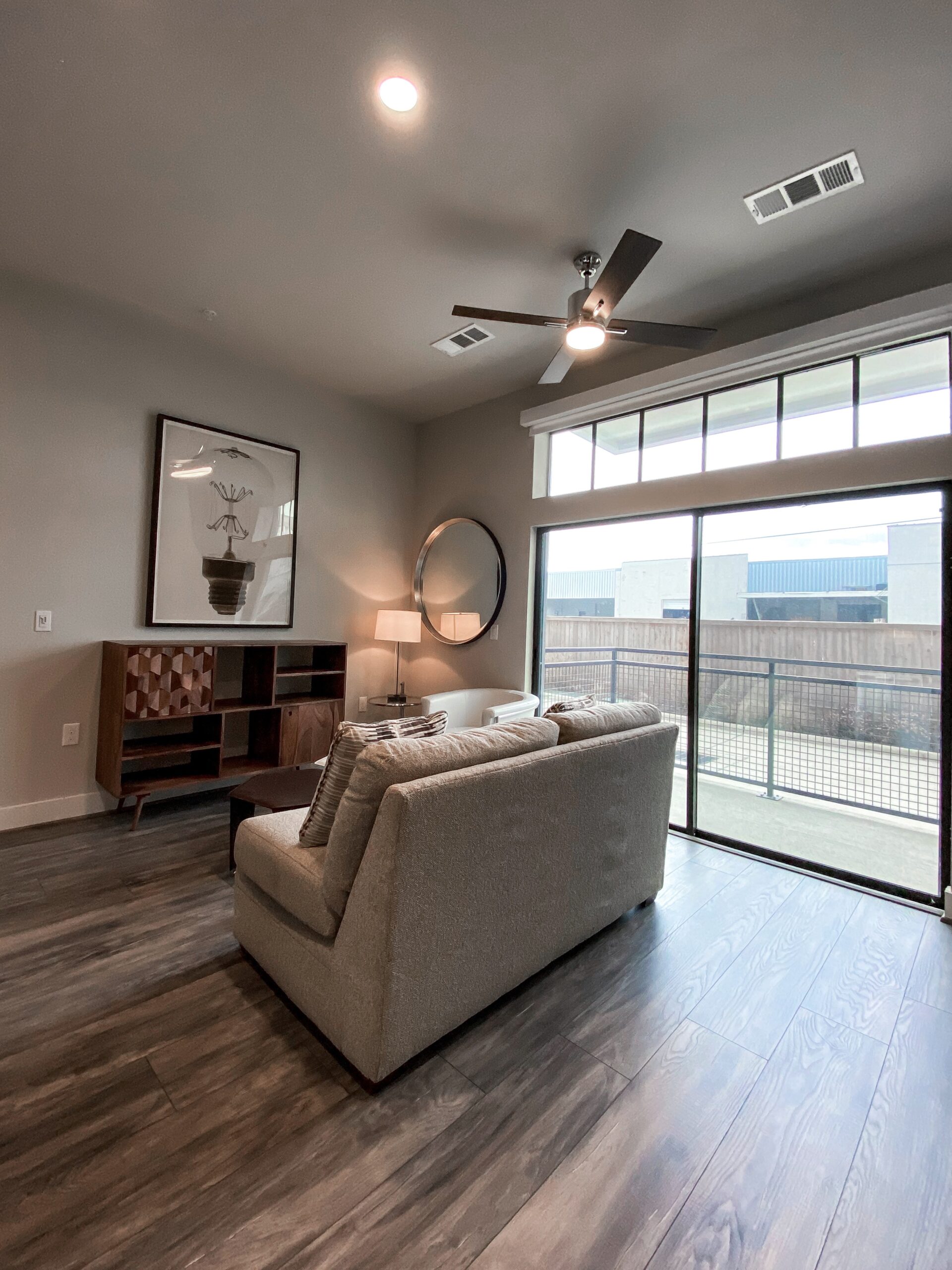 A modern living room features a light gray sofa with striped cushions, positioned near a large glass door that opens to a balcony. A round mirror hangs on the wall beside a framed artwork depicting a light bulb. A sleek floor lamp stands next to a small, round table, and a wooden cabinet with a geometric design is placed against the wall. Natural light fills the space, highlighting the contemporary decor.