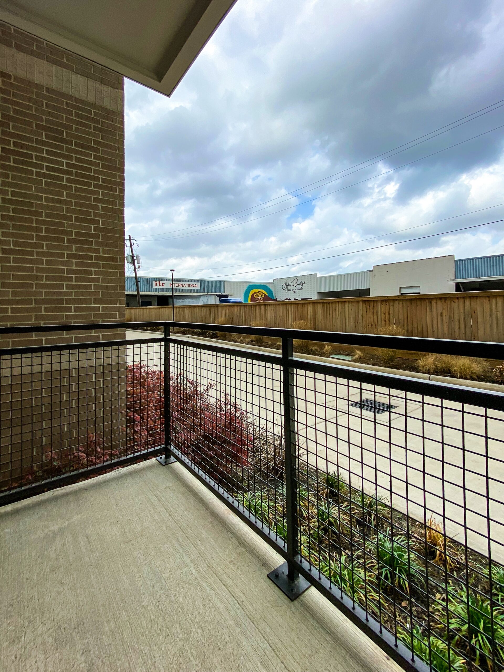 A view from a balcony shows a brick wall on the left and a metal railing with a grid pattern. Below the railing, there are some reddish plants. In the background, a street is visible with a wooden fence and several commercial buildings, including one with the sign "ITC INTERNATIONAL" and another featuring a colorful mural. The sky is partly cloudy, adding a soft light to the scene.