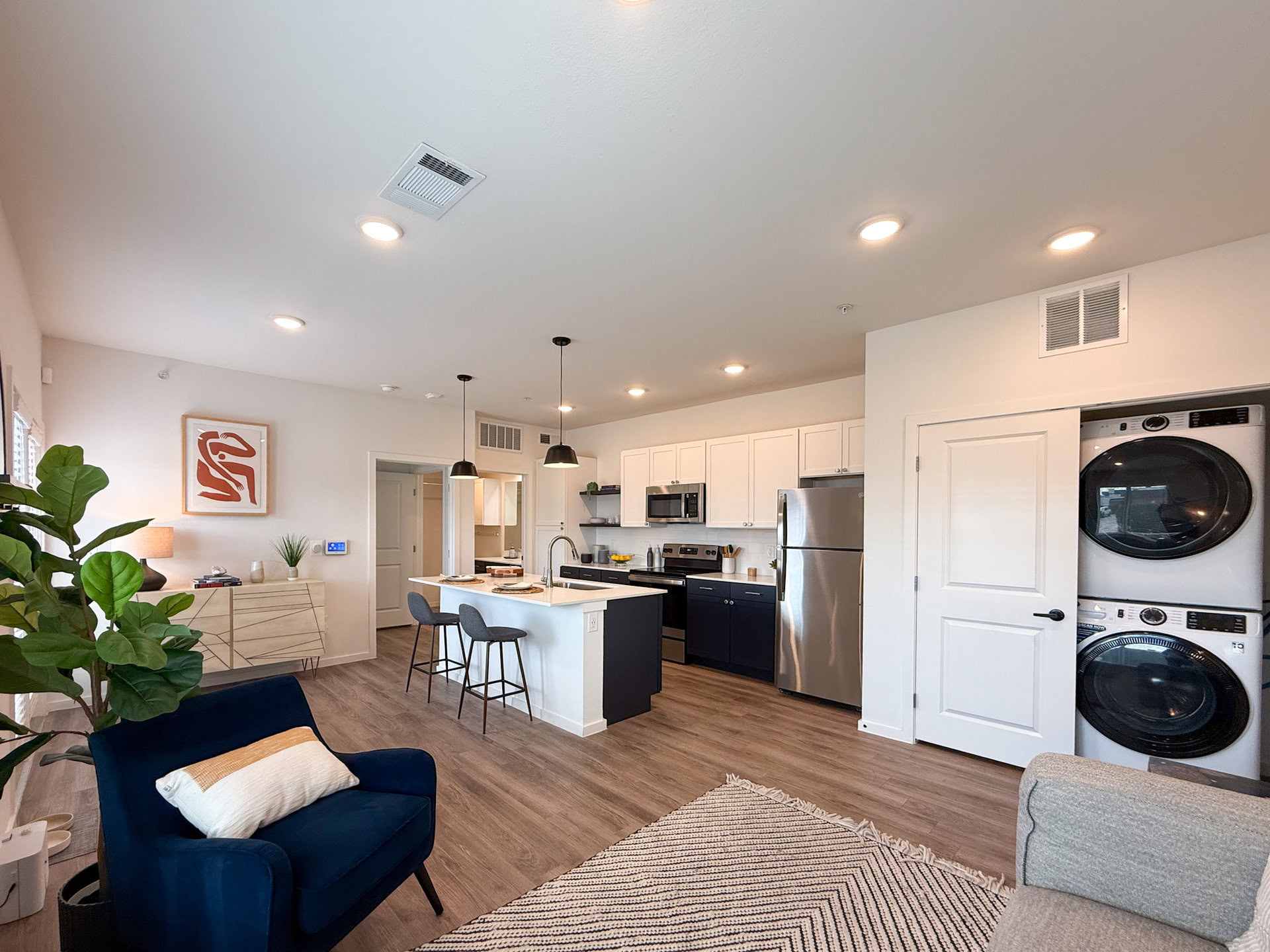 A modern kitchen and living area featuring an open layout. The kitchen has white cabinets and dark blue lower cabinets, with stainless steel appliances including a refrigerator and microwave. A large island with bar seating is positioned in the center. Two pendant lights hang above the island. To the side, there is a small dining area. A washer and dryer are stacked in a closet near the kitchen. The flooring is a light wood.