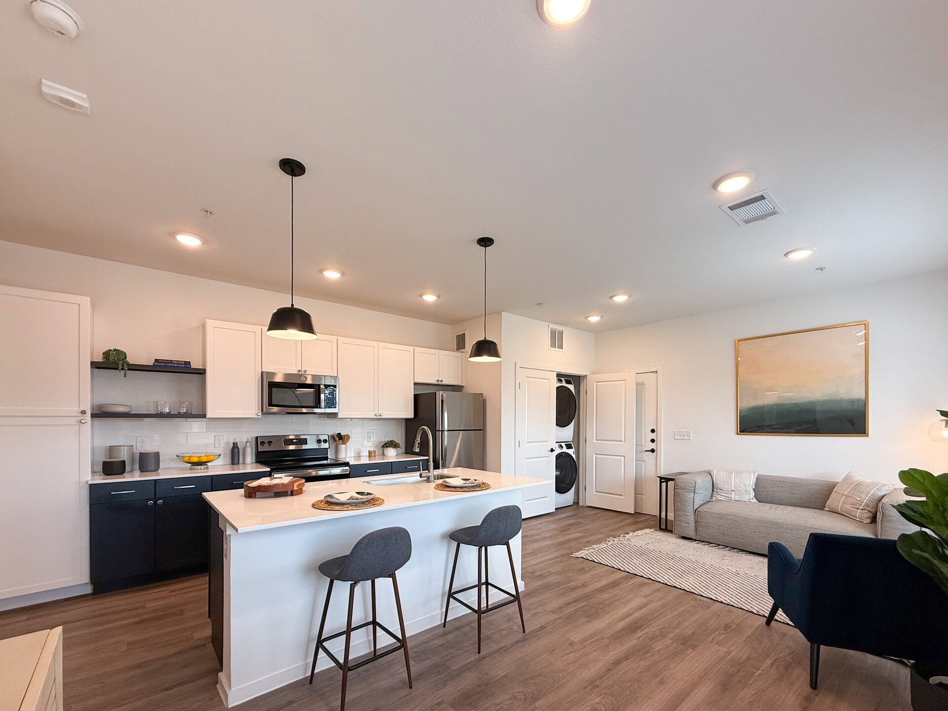 A modern kitchen and living area featuring an open layout. The kitchen has white cabinets and dark blue lower cabinets, with stainless steel appliances including a refrigerator and microwave. A large island with bar seating is positioned in the center. Two pendant lights hang above the island. To the side, there is a small dining area. A washer and dryer are stacked in a closet near the kitchen. The flooring is a light wood.