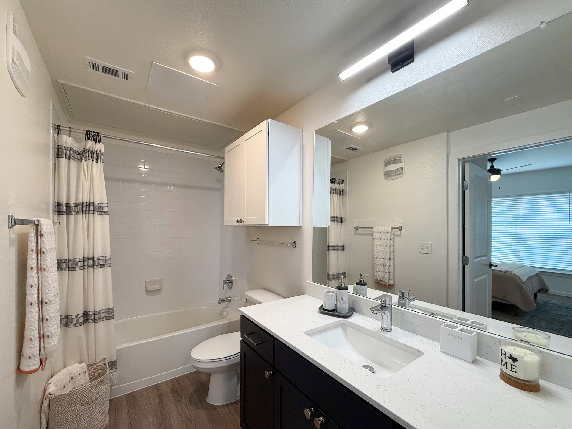 A modern bathroom features a white bathtub with a shower curtain, a sleek vanity with a white countertop, and white/dark cabinetry. The mirror above the sink reflects the space. The lighting is bright and contemporary, enhancing the clean aesthetic of the space.
