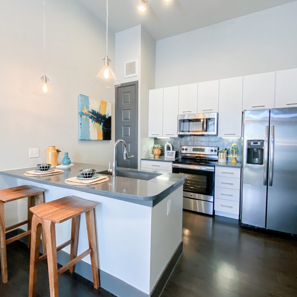 A modern kitchen features sleek white cabinetry and stainless steel appliances, including a refrigerator, oven, and microwave. A dark granite countertop extends into a small breakfast bar with two wooden stools. Above the bar, three pendant lights provide illumination. A colorful abstract painting hangs on the wall, complementing decorative items like a pitcher and a small plant on the counter. The flooring is dark, enhancing the contemporary aesthetic of the space.