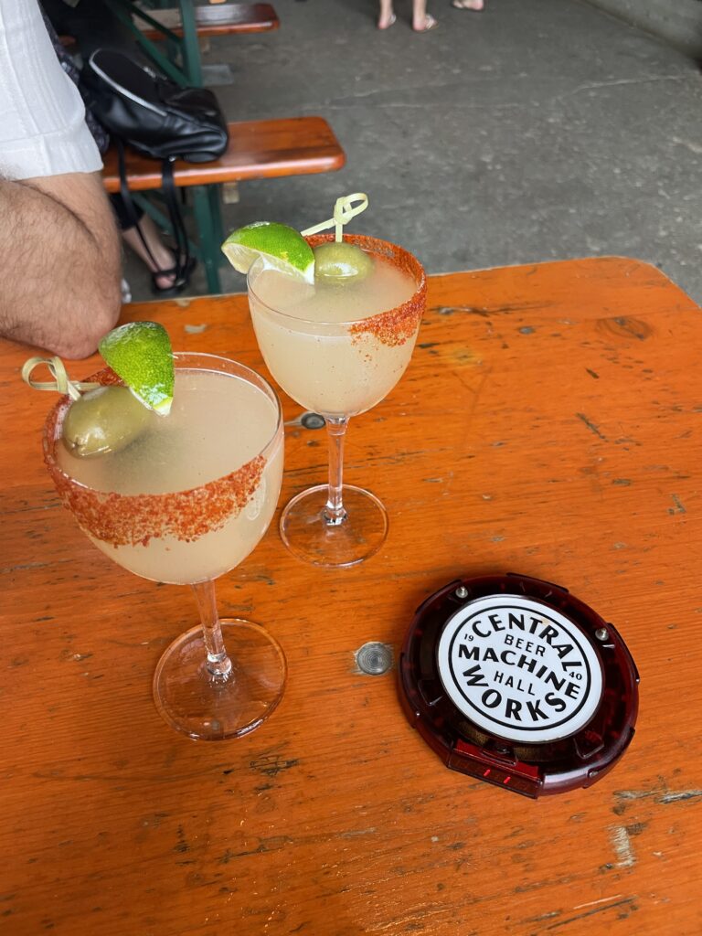 Two cocktails are displayed on a wooden orange table. Each drink is served in a clear glass with a rim coated in red seasoning. One cocktail features a green lime wedge and an olive as garnishes. The setting appears casual and inviting.