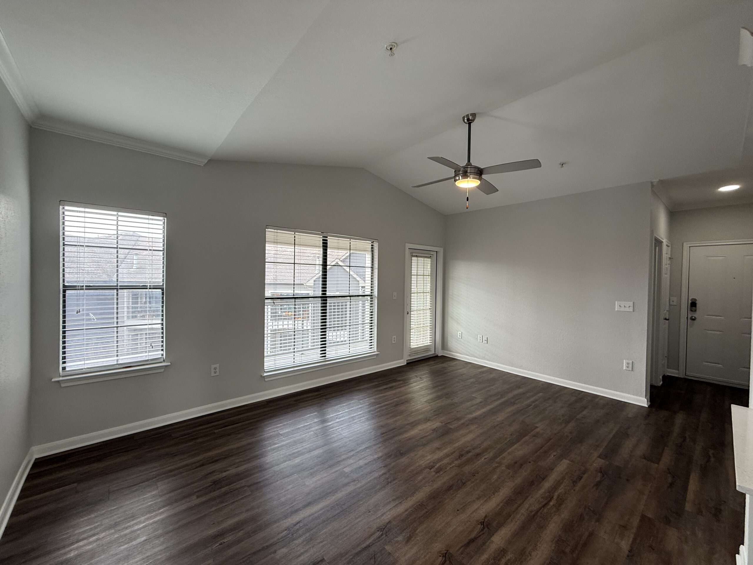 A spacious living area features light gray walls and dark wood laminate flooring. Two large windows with white blinds allow natural light to enter, while a ceiling fan with a light fixture hangs from the vaulted ceiling. A door with glass panels leads to an outdoor space, and a front door is visible on the right side of the image. The overall ambiance is bright and open, creating a welcoming atmosphere.