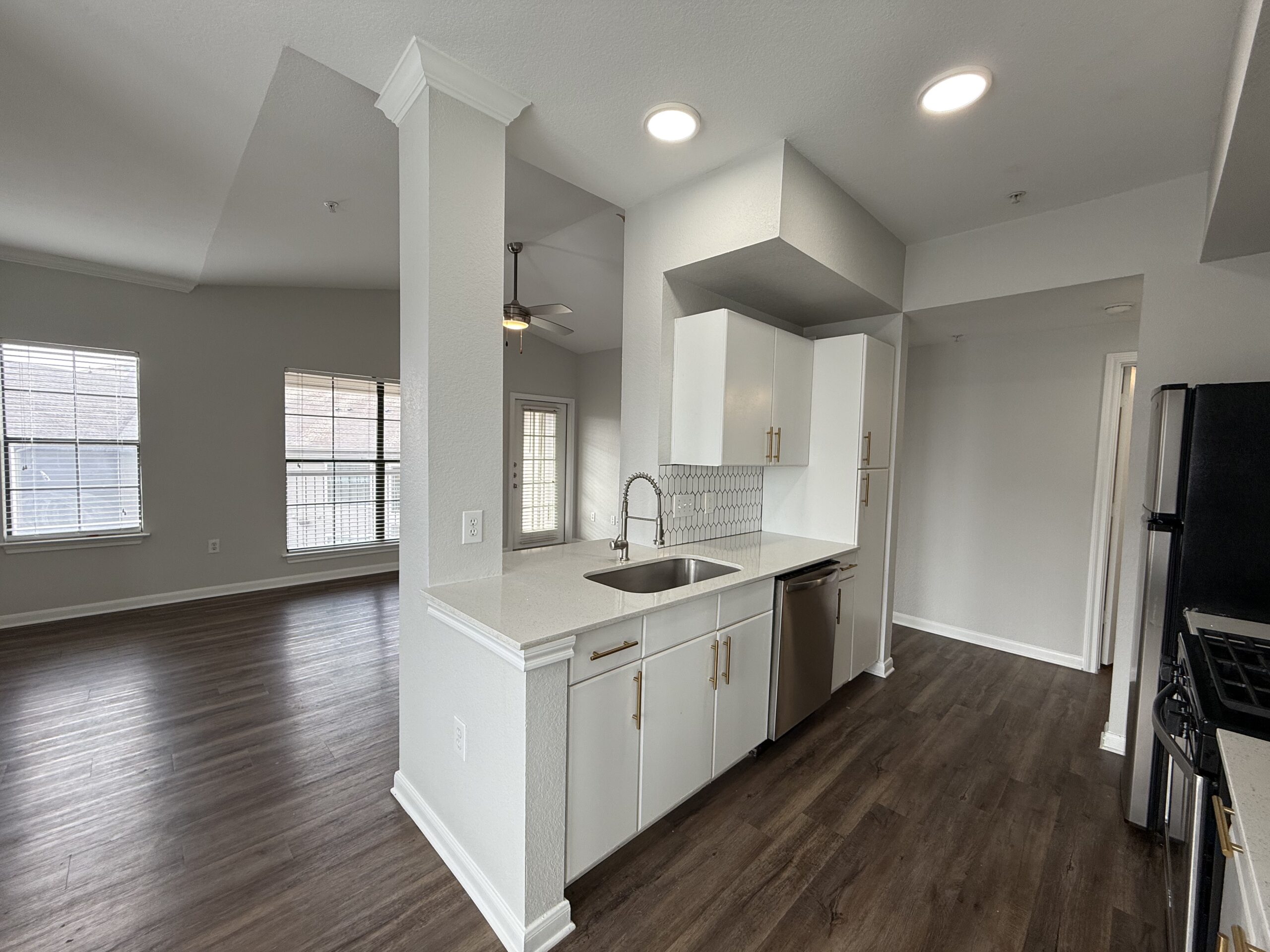 A modern kitchen area features white cabinetry with gold handles, a stainless steel sink, and a sleek countertop. The backsplash consists of hexagonal tiles. To the right, a black refrigerator and a gas stove are visible. The space opens into a well-lit living area with large windows adorned with blinds, showcasing a light-colored wall and wooden flooring throughout. A ceiling fan is mounted in the corner, adding to the contemporary feel.