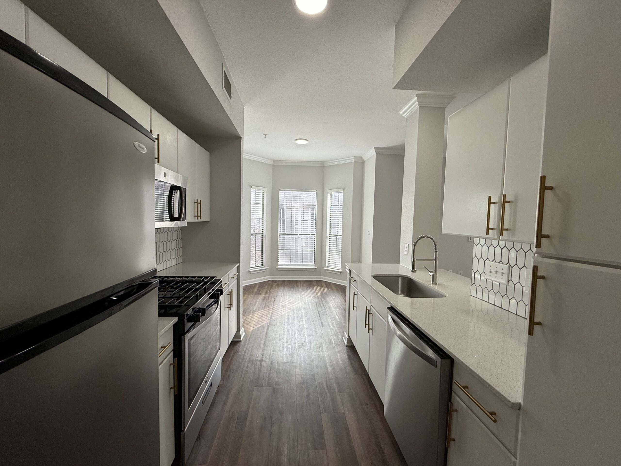 A modern kitchen features white cabinetry with gold handles, a stainless steel sink, and a countertop with a light-colored stone finish. Appliances include a black refrigerator, a gas stove, and a microwave. The backsplash consists of hexagonal tiles. Natural light floods the space through multiple windows in a bay area, highlighting the wood-like flooring. The overall design is sleek and contemporary.