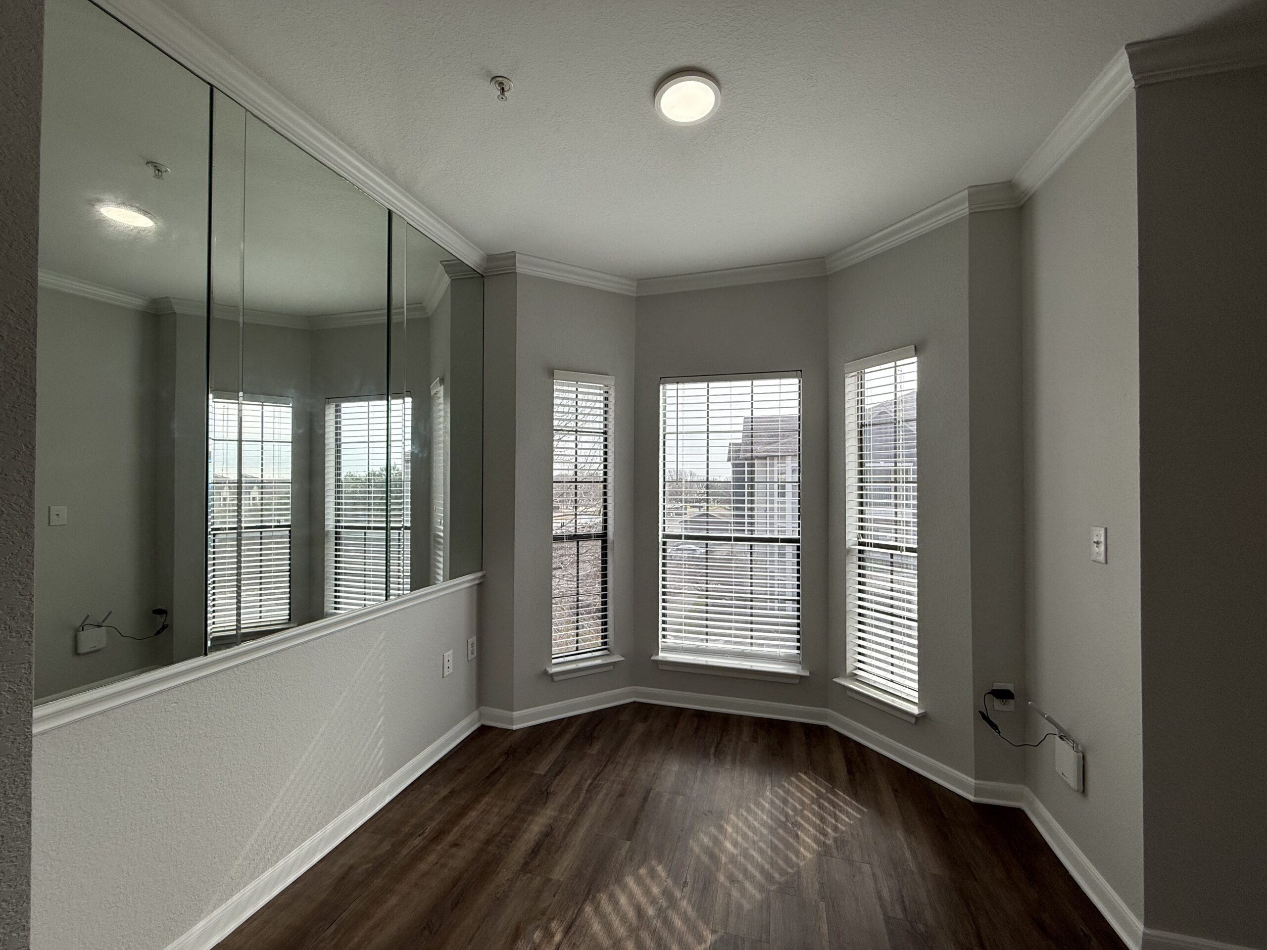 A corner room features large windows with white blinds, allowing natural light to fill the space. The walls are painted in a light gray color, and there is a mirrored wall that reflects the room's brightness. The flooring is a warm, dark wood, and there is a small electrical outlet visible on the wall. The ceiling has a simple crown molding, enhancing the room's elegant appearance.