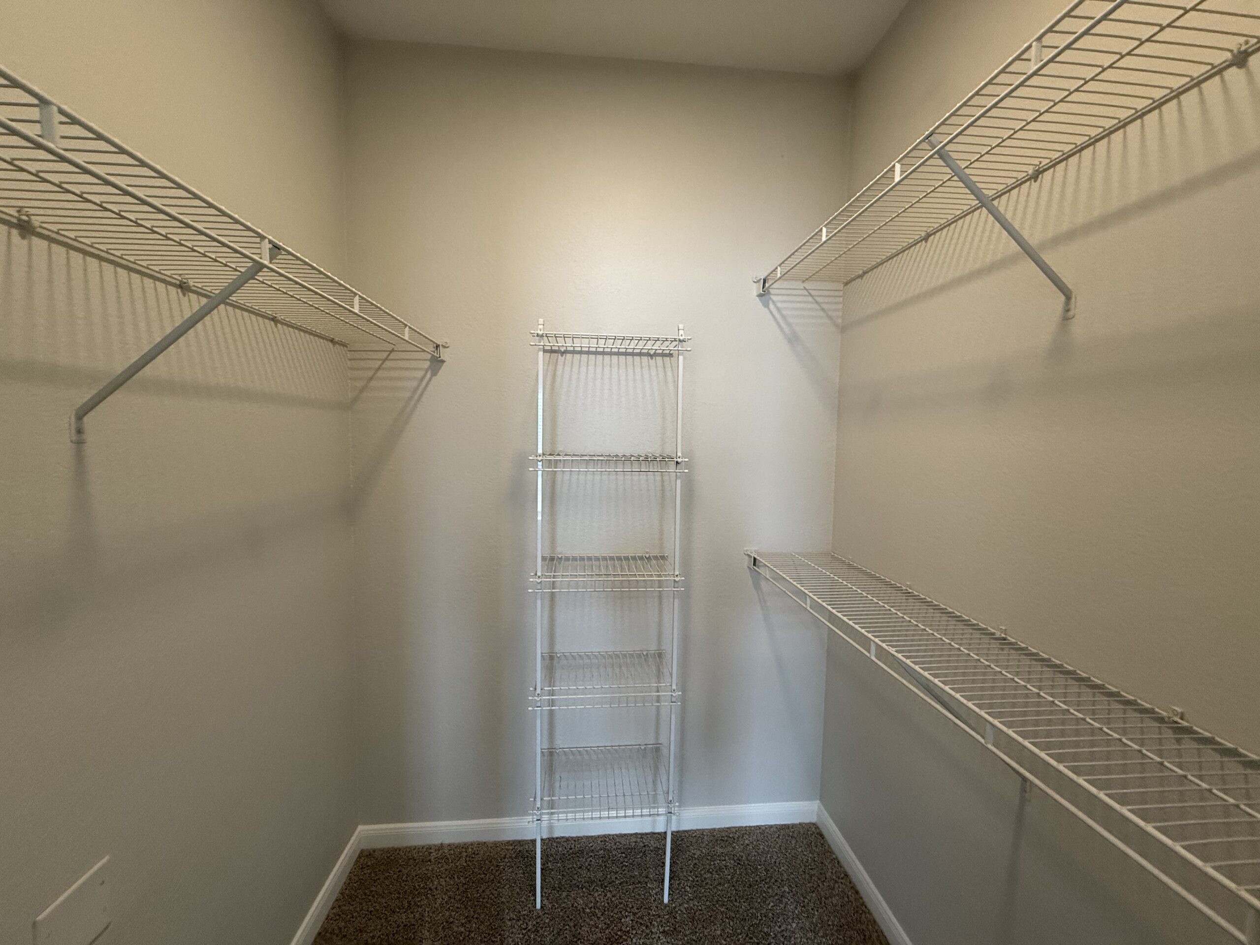 A spacious closet features multiple wire shelving units. Two long shelves run along the upper walls, while a tall, narrow shelving unit with five tiers stands in the center. The walls are painted a light color, creating a bright and open feel. The floor is clear, emphasizing the organized storage potential of the space.
