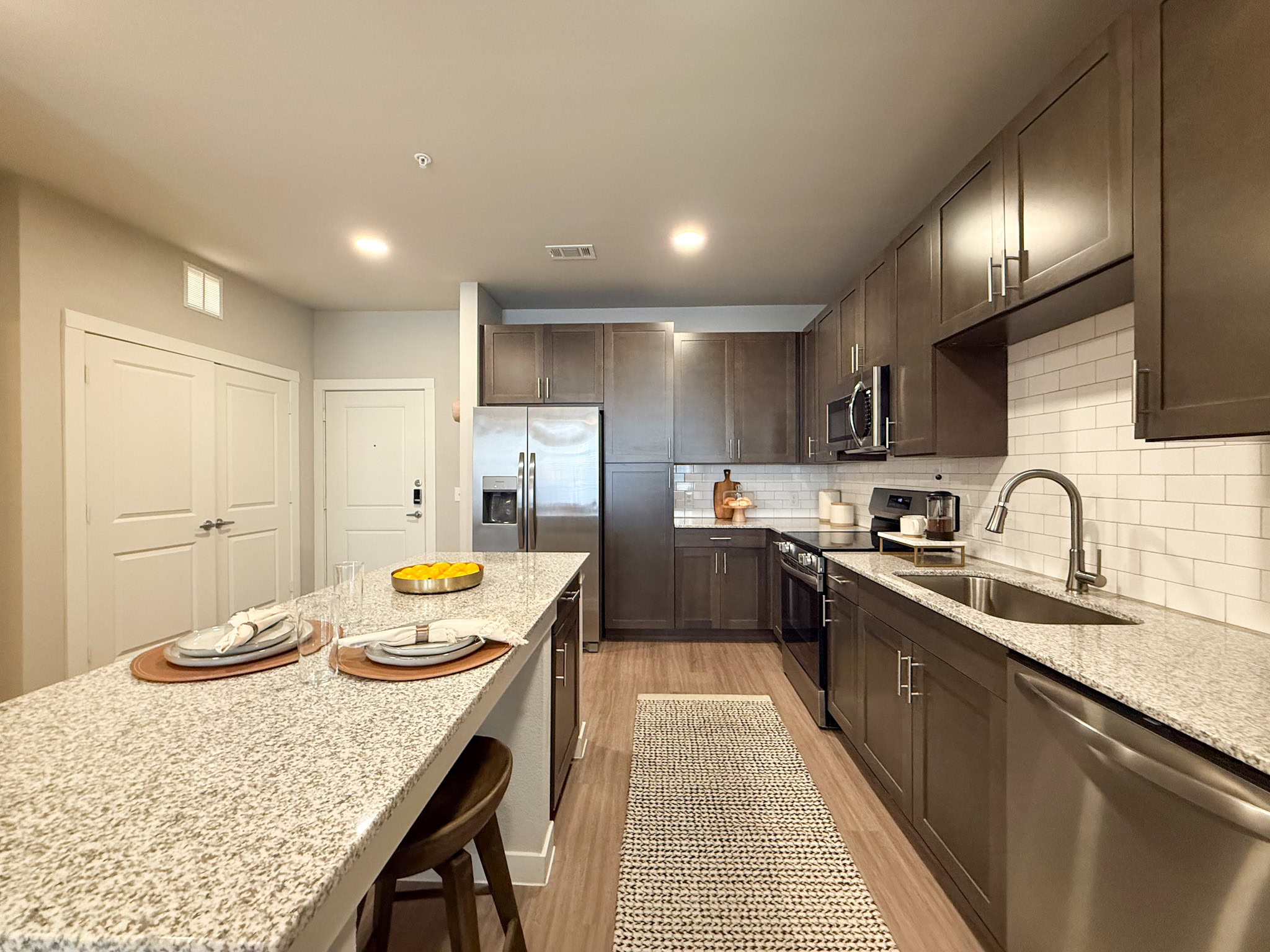 A modern kitchen featuring dark wood cabinetry and stainless steel appliances. The countertop is made of light-colored granite, complemented by a white subway tile backsplash. A large refrigerator. There are two bar stools at the island. The space is well-lit with overhead lighting and has a clean, contemporary design.