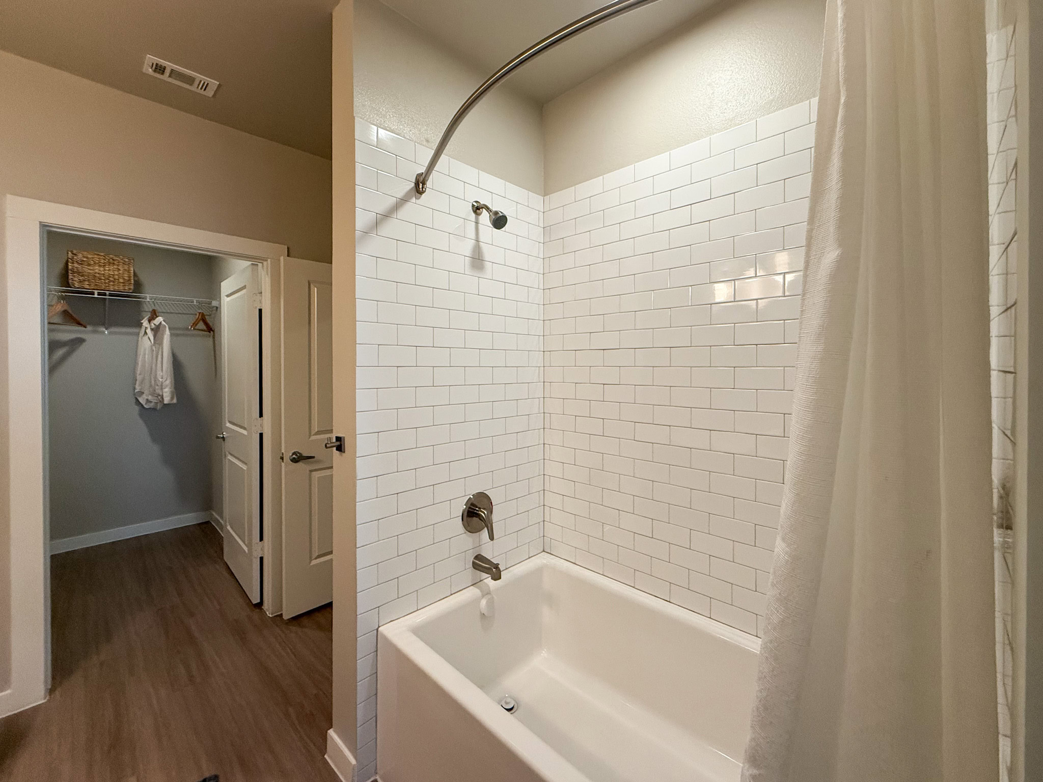 A modern bathroom features a white tiled shower area with a curved shower rod and a white curtain. The bathtub is white and has a sleek faucet and showerhead. The flooring is a light wood finish.