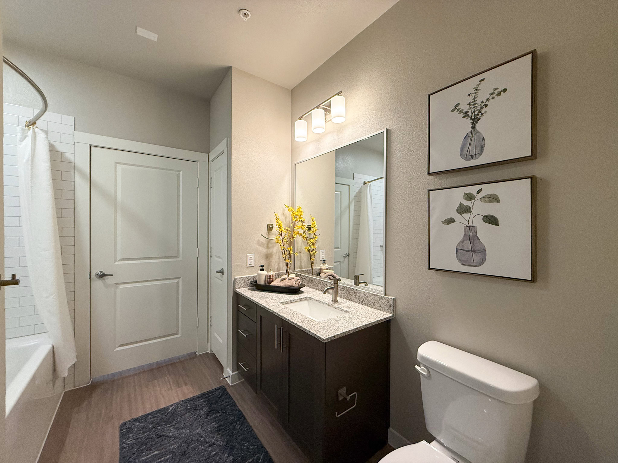 A modern bathroom features a sleek vanity with a granite countertop and dark wood cabinetry. Above the sink, a large mirror is illuminated by three cylindrical light fixtures. A white shower curtain partially conceals a bathtub with white subway tile, and a white toilet is positioned nearby. The flooring is a warm wood tone, contributing to the contemporary aesthetic.