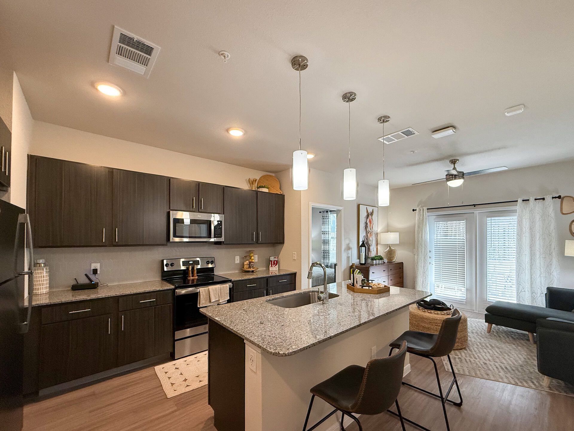 A modern kitchen features dark wood cabinetry and stainless steel appliances, including a microwave and oven. A large granite countertop with an undermount sink serves as a central workspace. Large windows with blinds allow natural light to fill the room. A small dining area or work from home station near the kitchen and living room.