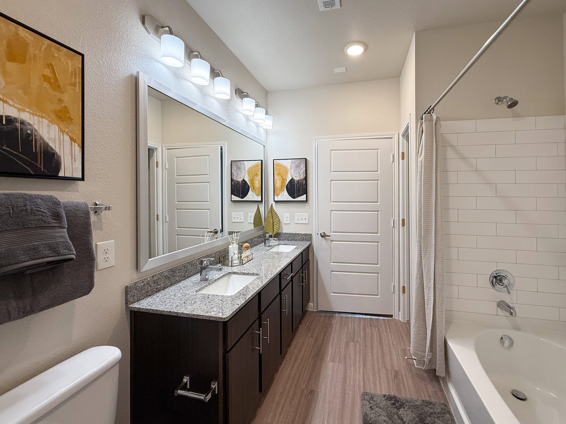 A modern bathroom features a double vanity with a granite countertop, two sinks, and stylish faucets. Above the vanity, a large mirror reflects the space, illuminated by contemporary light fixtures. The shower area is enclosed with white subway tiles and a curtain. The flooring is a warm wood-like laminate.