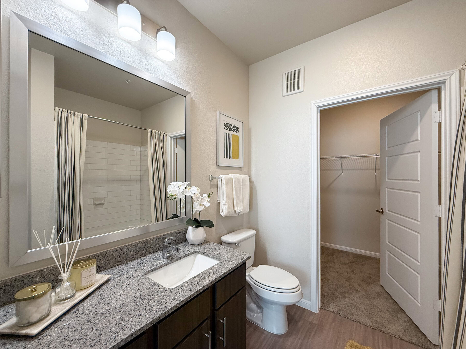 A modern bathroom features a granite countertop with a sink and faucet. A large mirror hangs above the sink, reflecting the space. A shower area, partially concealed by striped curtains, showcases white subway tiles, and the walls are painted in a soft neutral tone, enhancing the bright and clean aesthetic.