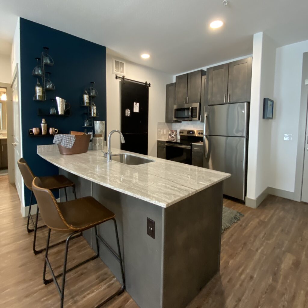 A modern kitchen features a large, light-colored granite countertop with a built-in sink. Three brown leather bar stools with metal frames are positioned at the counter. The back wall is painted a deep blue and showcases glass shelves holding various kitchen items. To the right, there are dark wood cabinets and stainless steel appliances, including a refrigerator, microwave, and oven. A black sliding barn door is visible, leading to another area of the space. The flooring is a warm wood tone, enhancing the contemporary feel of the kitchen.