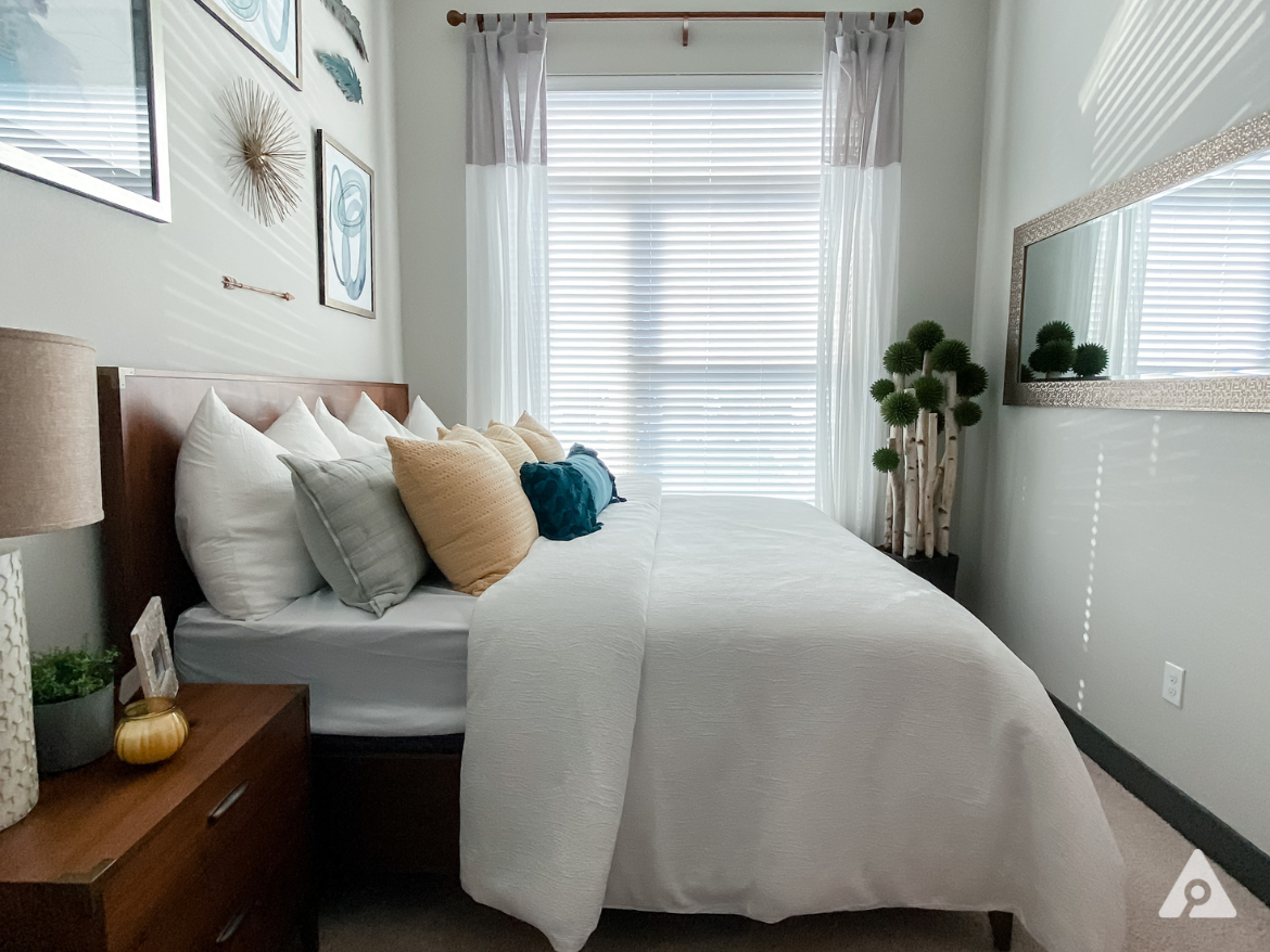 A cozy bedroom features a queen-sized bed with a white quilt and an array of decorative pillows in various colors, including white, gray, and teal. The headboard is wooden, and the bed is positioned against a wall adorned with framed artwork and decorative elements. Natural light streams through a large window covered with sheer curtains, casting soft shadows. To the side, a nightstand holds a lamp and a small plant, while a mirror reflects the room's decor. A decorative plant arrangement adds a touch of greenery to the space.