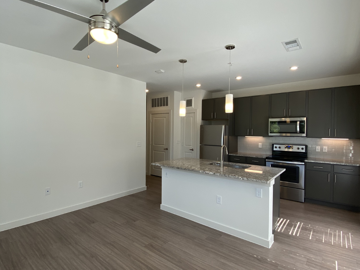 A modern kitchen and living area featuring an open layout. The kitchen has dark cabinetry, stainless steel appliances, and a large granite island with a sink. Pendant lights hang above the island, and there is a ceiling fan in the living area. The flooring is a light wood laminate, and the walls are painted a soft white. Natural light streams in from a window, creating a bright and inviting atmosphere.