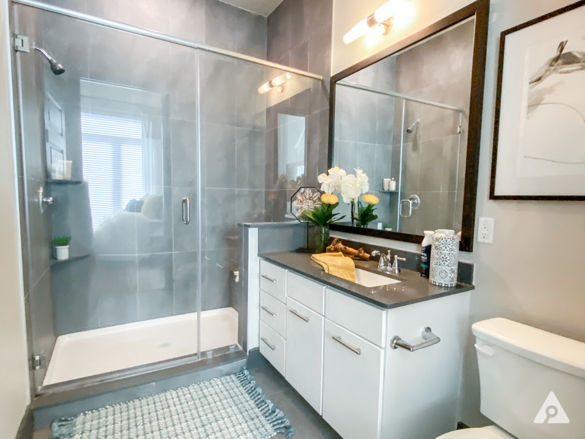 A modern bathroom features a spacious glass shower with a sleek design. The walls are tiled in a light gray, complemented by a dark gray countertop. A white vanity with multiple drawers holds a sink, and decorative items include a yellow towel and a small potted plant. Above the sink, a large mirror is framed in black, reflecting the room's light. A white toilet is positioned beside the vanity, and a textured rug lies on the floor, adding warmth to the space. Natural light filters in through a window, enhancing the overall ambiance.
