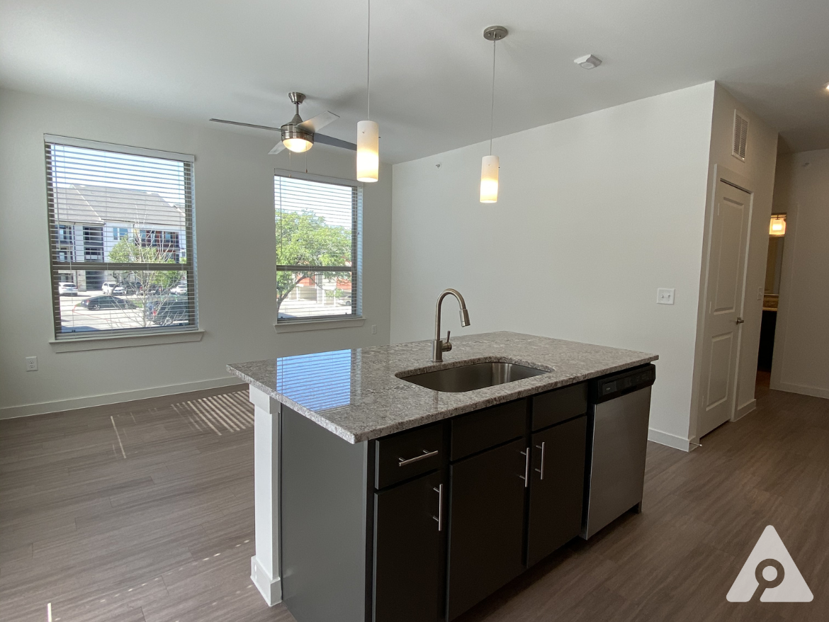 A modern kitchen area features a central island with a granite countertop and a sink. Dark cabinetry complements the stainless steel dishwasher. Two large windows with blinds allow natural light to fill the space, revealing a view of the outside. A ceiling fan and a pendant light provide additional illumination. The flooring is a light wood laminate, enhancing the contemporary feel of the room. A doorway in the background leads to another area of the apartment.