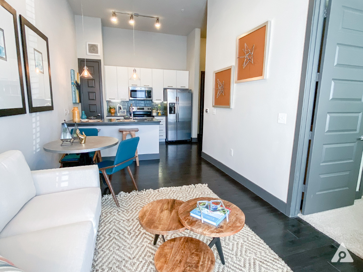 A modern living space features a light-colored sofa and a round table with teal chairs. The kitchen area is visible in the background, showcasing white cabinetry, stainless steel appliances, and pendant lighting. A textured area rug lies beneath two wooden coffee tables. On the walls, there are framed artworks, and a door leads to another room. The flooring is dark wood, enhancing the contemporary aesthetic.