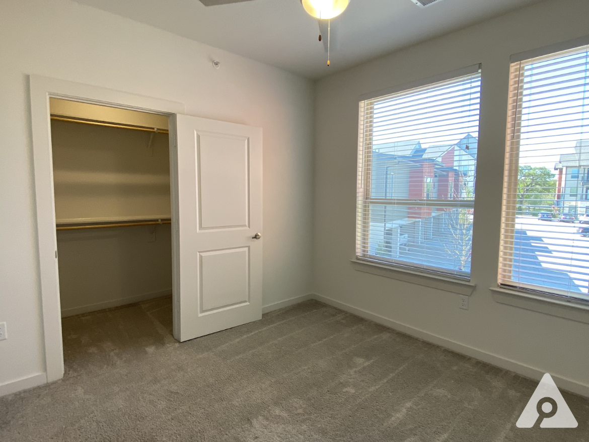 A well-lit bedroom features two large windows with blinds, allowing natural light to enter. The walls are painted in a light color, and the floor is covered with soft, beige carpet. On one side, there is a closet with a white door, which is slightly ajar, revealing a shelf and hanging space inside. The ceiling has a ceiling fan with a light fixture. The overall atmosphere is bright and inviting.