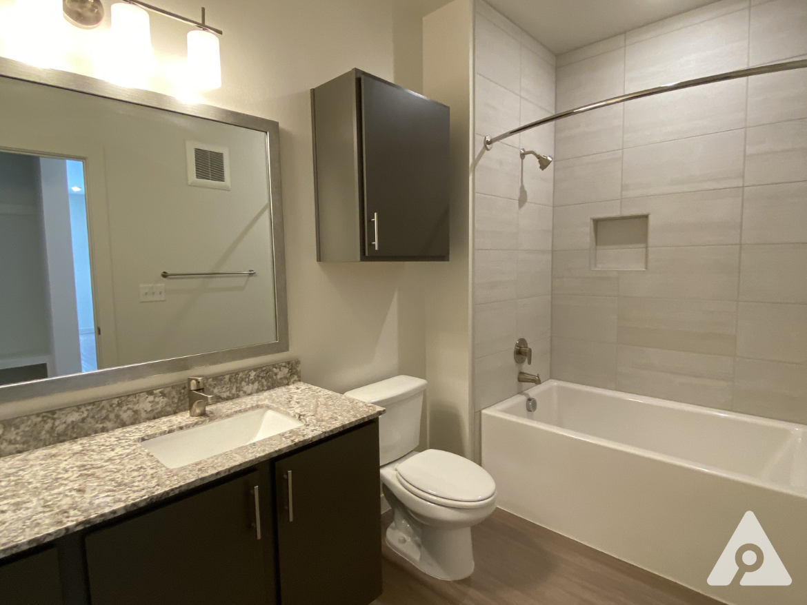 A modern bathroom features a sleek design with light-colored tile walls and a curved shower rod. The space includes a white bathtub with a built-in niche for toiletries, a toilet, and a dark cabinetry unit beneath a granite countertop with an integrated sink. Above the sink, a large rectangular mirror is framed in silver, and three light fixtures provide illumination. The flooring is a warm wood-like material, enhancing the contemporary feel of the room.