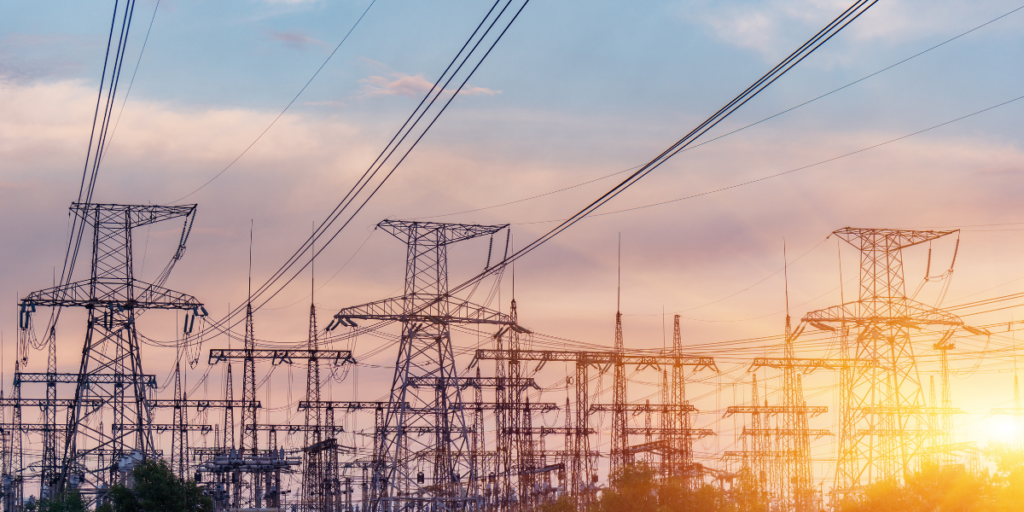 A series of tall electrical transmission towers are silhouetted against a colorful sunset sky. The towers are interconnected by numerous power lines, creating a complex network. The background features soft clouds illuminated by the warm hues of the setting sun, while the foreground includes some greenery at the base of the towers.