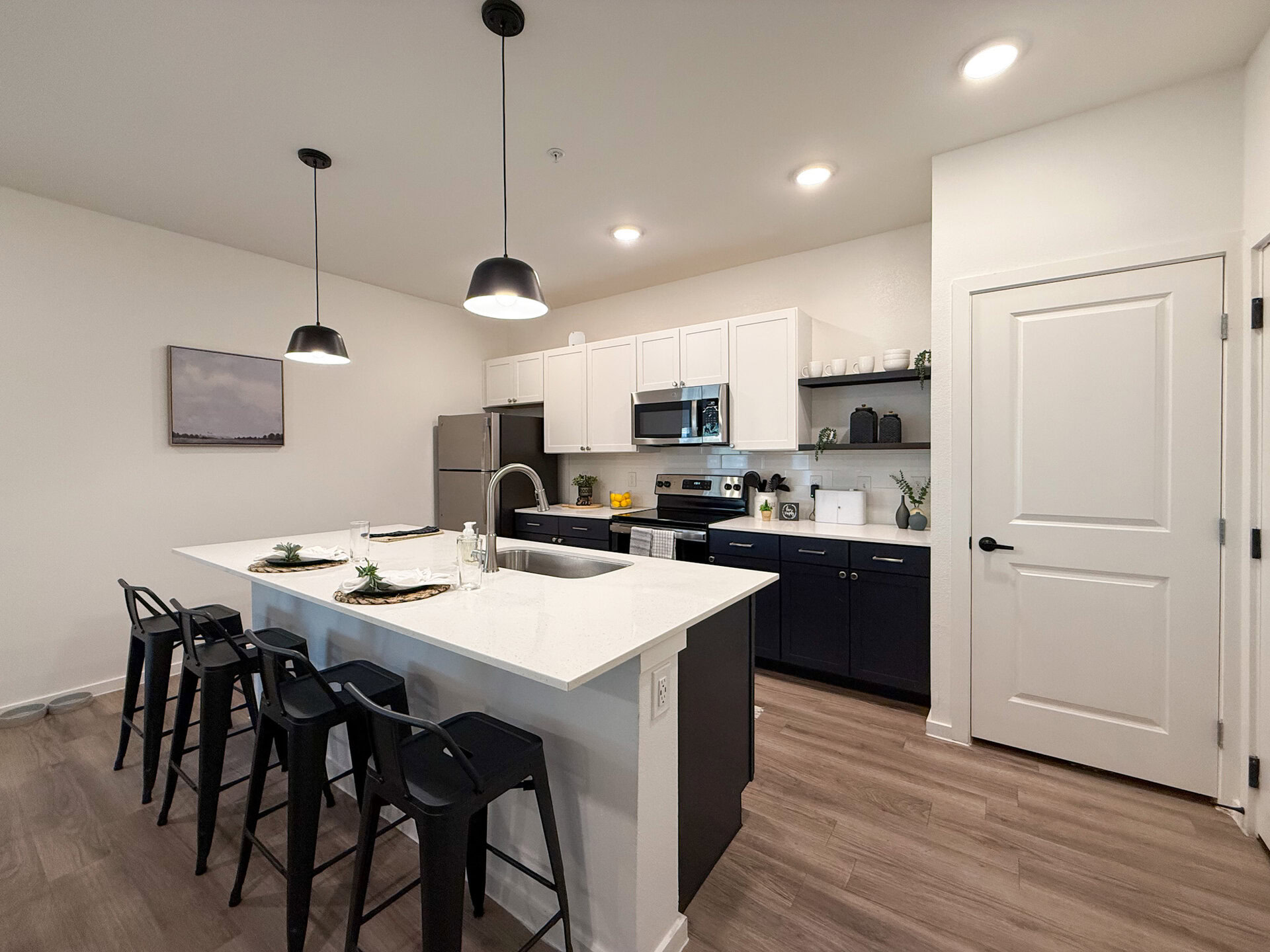 A modern kitchen features a large island with a white countertop and black bar stools. Above the island, two black pendant lights hang. The kitchen cabinetry is a mix of white upper cabinets and dark blue lower cabinets, with stainless steel appliances, including a refrigerator and an oven. A small shelf displays decorative items and plants. The flooring is a light wood finish giving modern finishes.