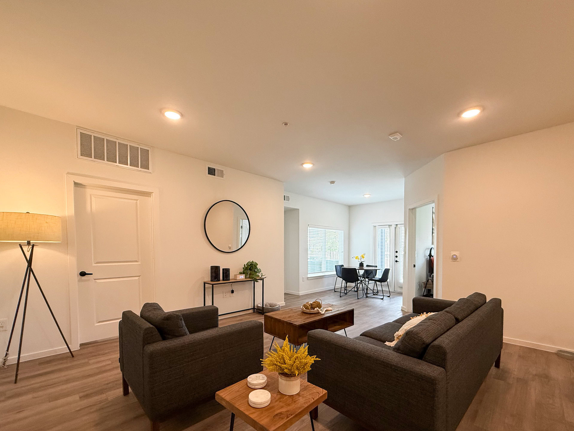 A modern living room features two gray sofas. In the background, a dining area is visible with a small table and four chairs, illuminated by natural light from a window.
