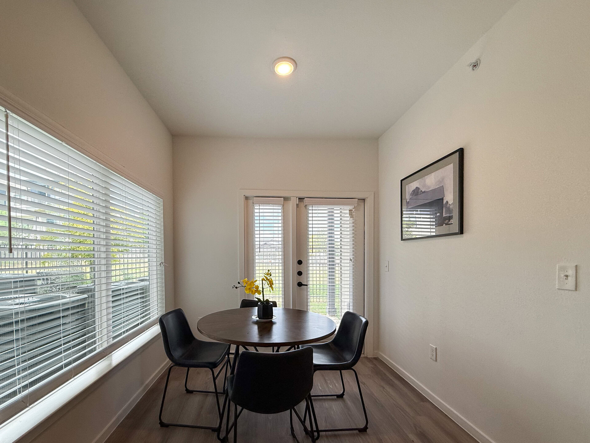 A small dining area features natural light streaming in through a large window with white blinds, and a pair of glass doors leads outside. The flooring is light-colored, enhancing the bright and airy atmosphere.