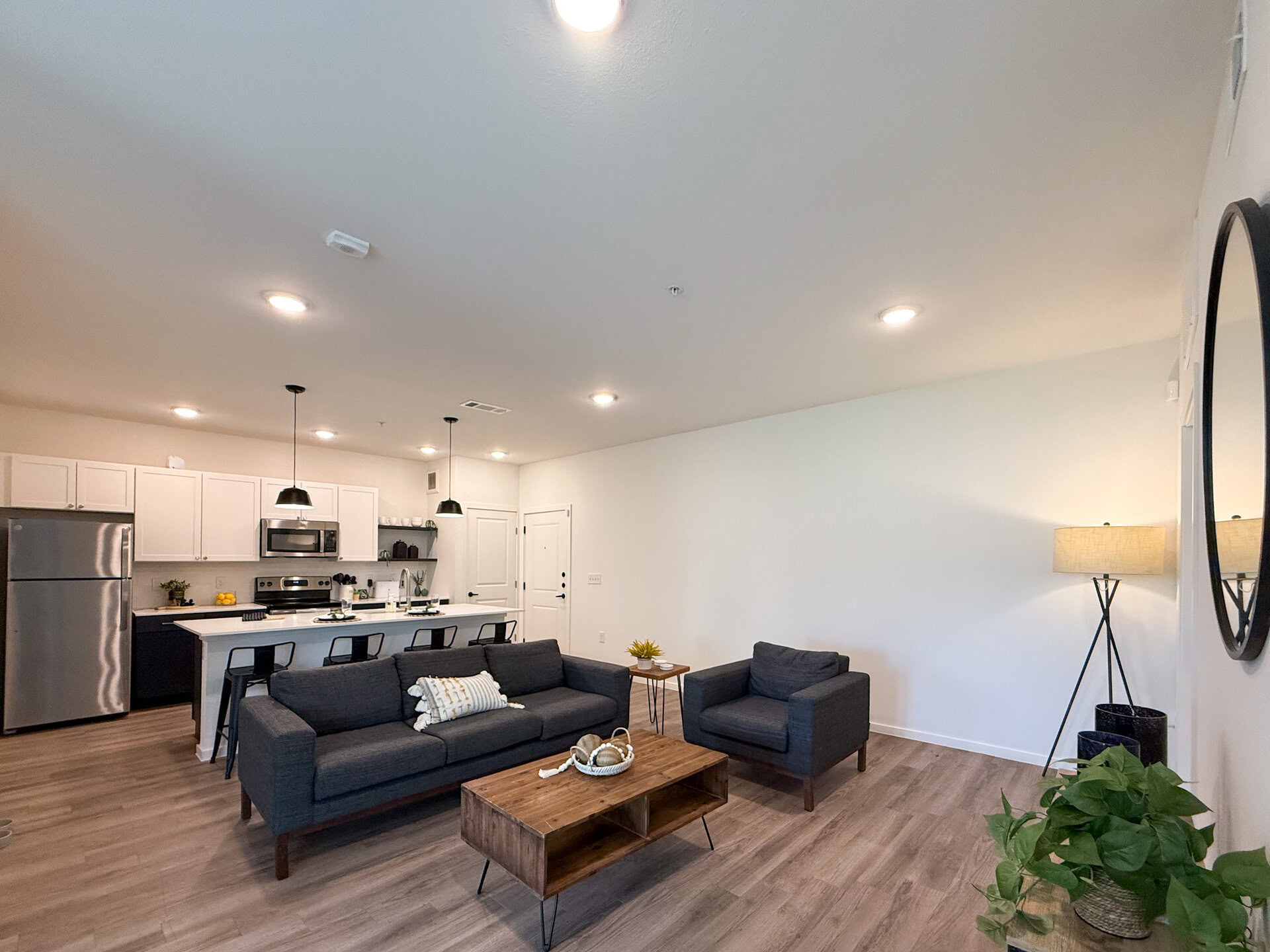 A modern living space features an open layout with a kitchen and living area. The kitchen has white cabinetry, stainless steel appliances, and a quartz countertop with bar seating. The living area includes a dark gray sofa & the flooring is light wood.