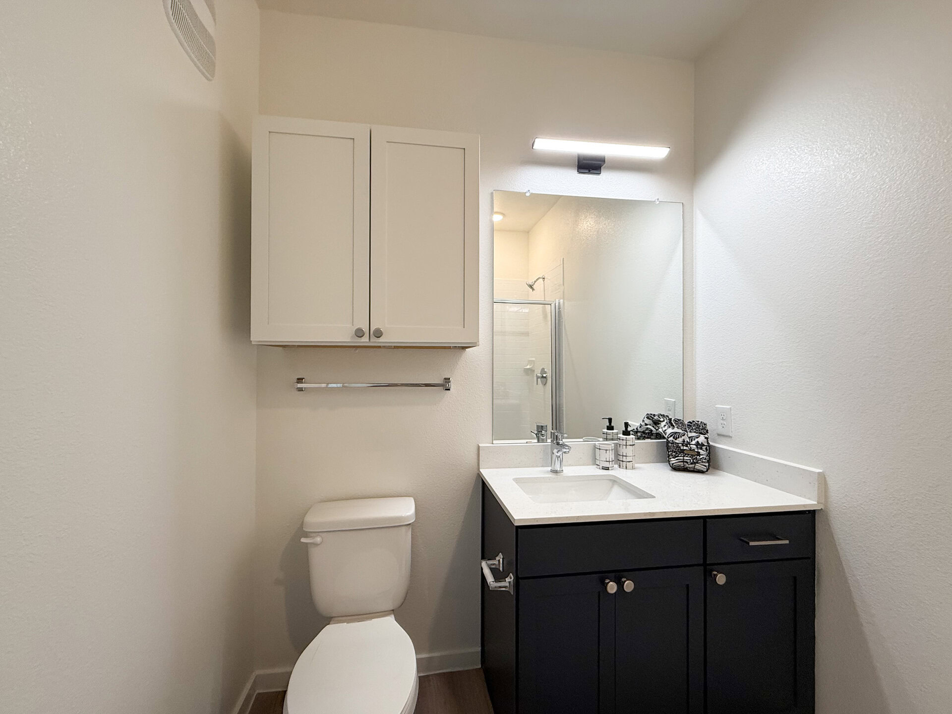 A modern bathroom features a white toilet and a dark cabinetry vanity with a quartz countertop. Above the vanity, there are two white cabinets for storage. In the back, a standing shower with white subway tiles.