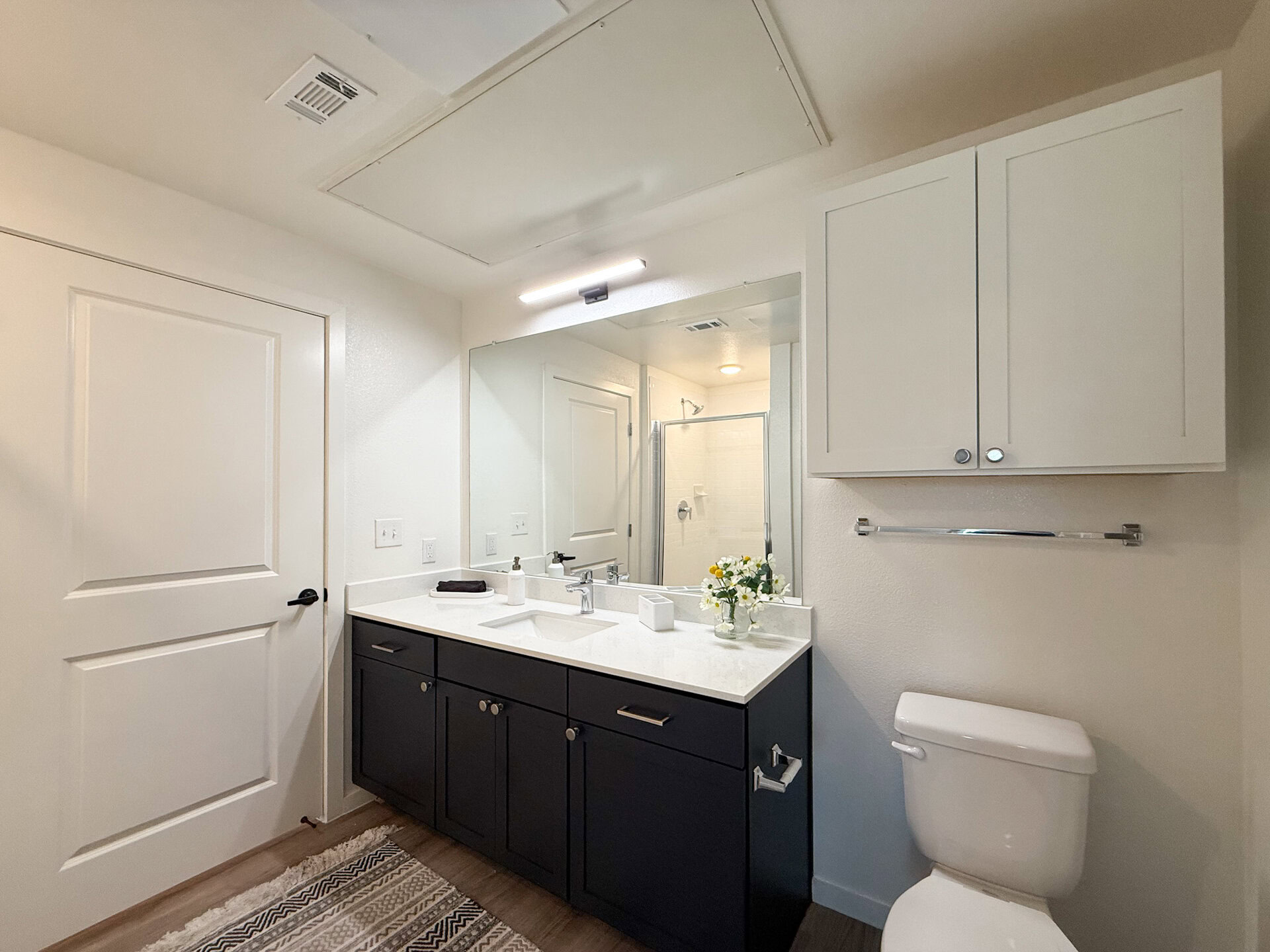 A modern bathroom features a double vanity, a white toilet, and a dark cabinetry vanity with a quartz countertop. Above the vanity, there are two white cabinets for storage. In the back, a standing shower with white subway tiles.