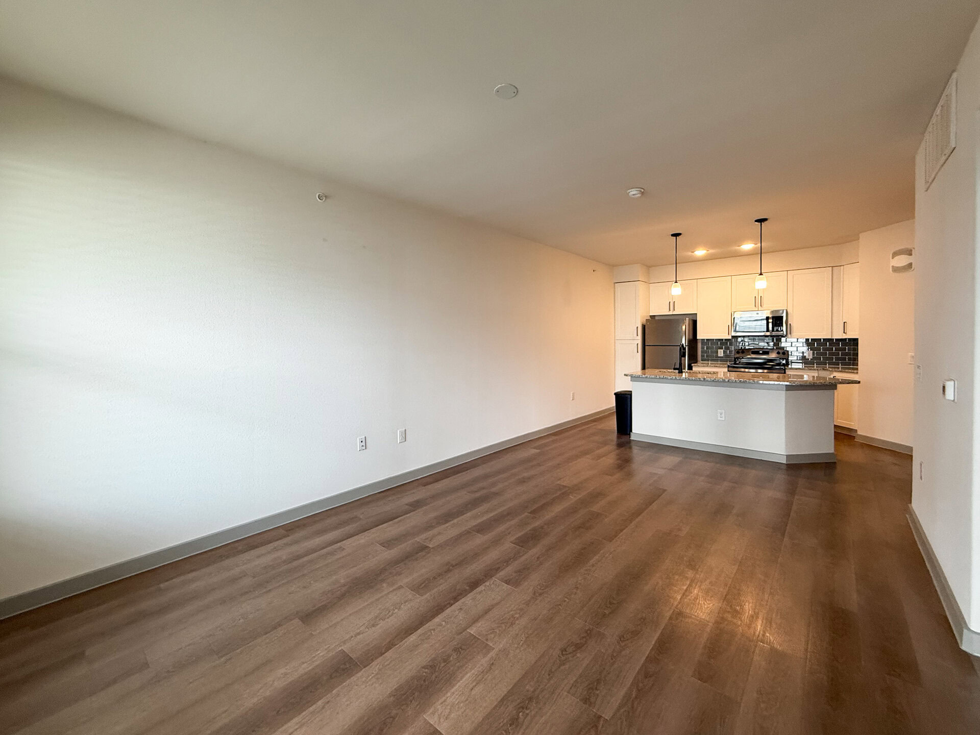 A spacious, modern living area features light-colored walls and a warm wood-like floor. To the right, a kitchen area is visible, showcasing white cabinetry, a stainless steel refrigerator, and a stove with a backsplash of dark tiles. Two pendant lights hang above the kitchen island, which has a granite countertop. The space is well-lit, with natural light coming from a window.