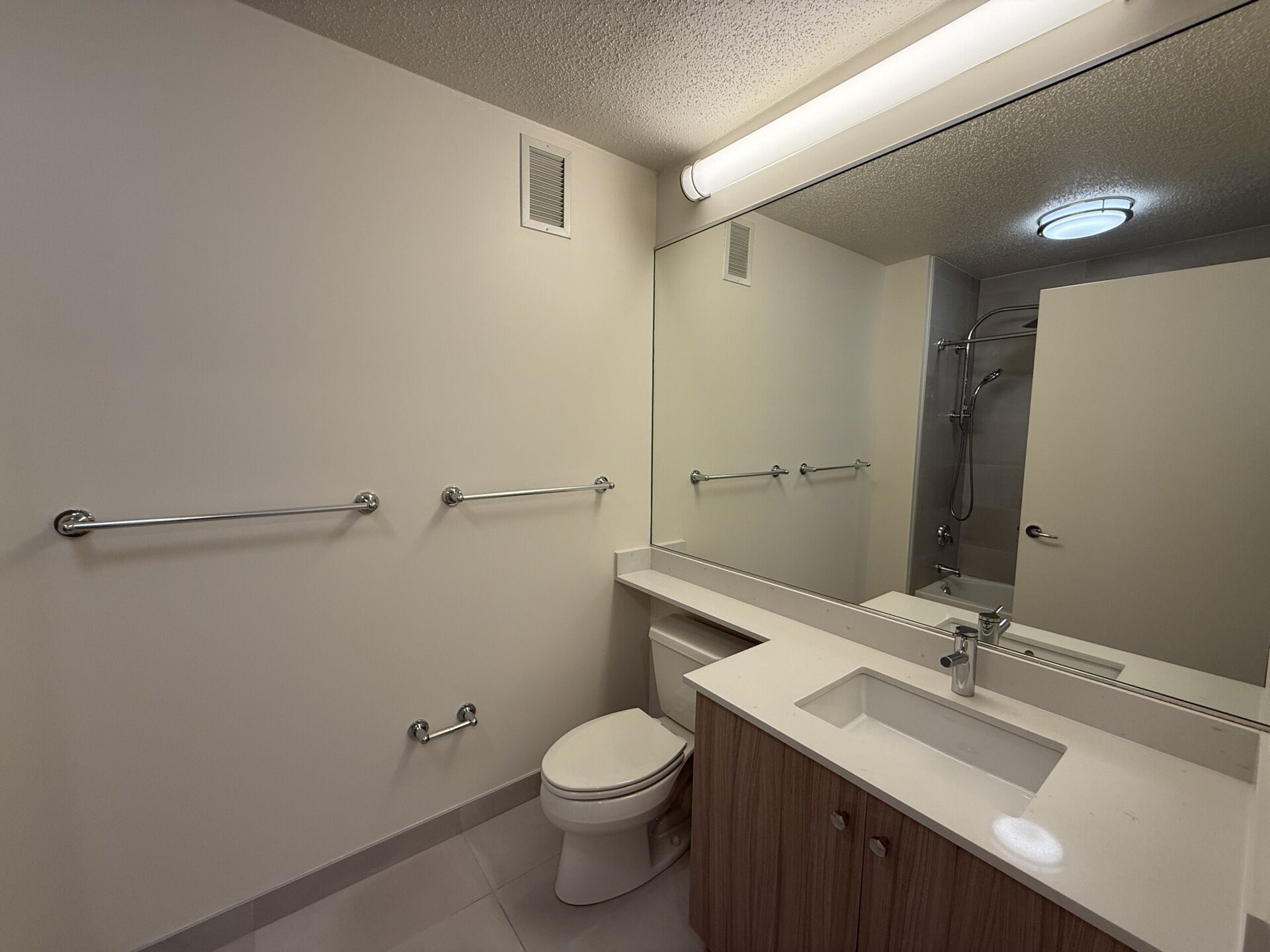 A modern bathroom features a clean, minimalist design. The space includes a white toilet and a wooden vanity with a rectangular sink. Above the sink, a large mirror reflects the room. On the wall, there are several chrome grab bars, and a shower area is visible in the background, equipped with a handheld showerhead. The lighting is provided by a ceiling fixture and a strip light above the mirror, creating a bright atmosphere. The walls are painted in a light color, enhancing the spacious feel of the room.
