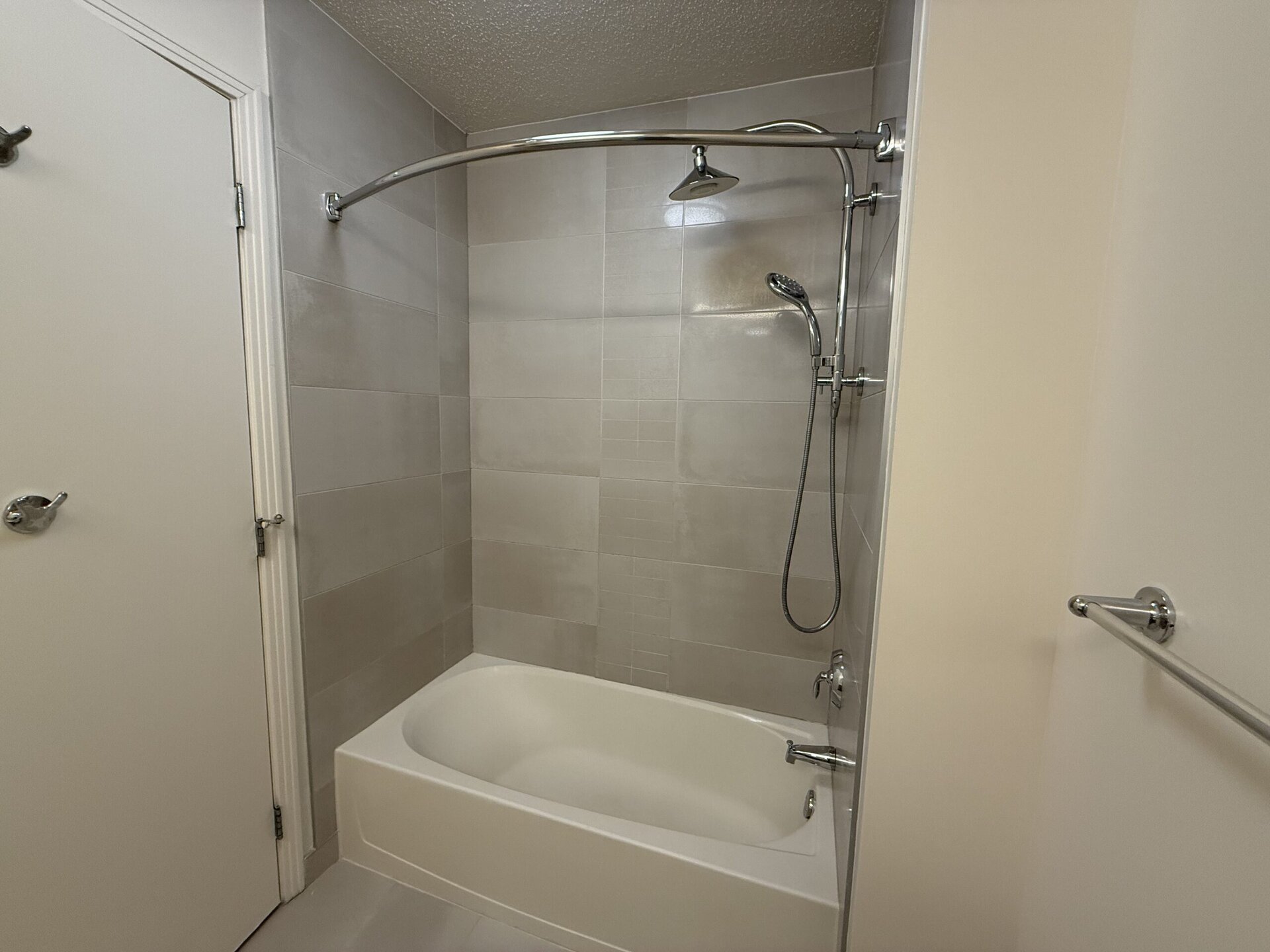 A modern bathroom features a bathtub with a showerhead mounted above. The walls are tiled in light gray, creating a clean and contemporary look. A chrome shower rod arches over the tub, and there is a handheld shower attachment on the wall. A towel bar is visible on the right side, and a white door is partially open on the left. The overall design is minimalistic and bright.