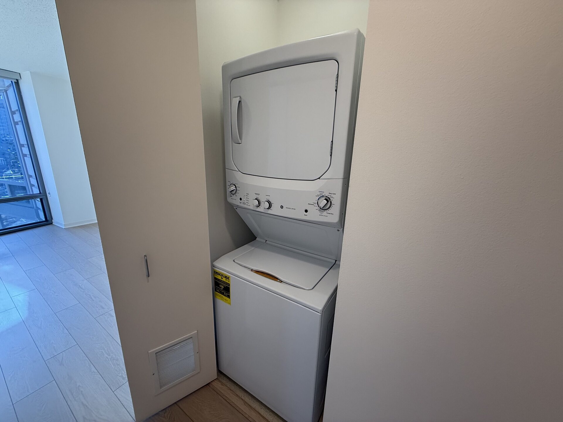 A stacked washer and dryer unit is positioned in a narrow space, partially obscured by a wall. The washer is at the bottom, featuring a top-loading design, while the dryer is above it with a front-loading door. Both appliances are white, and the control panels include various dials and settings. A small warning label is visible on the washer. The background shows a bright, airy room with large windows and light-colored flooring.
