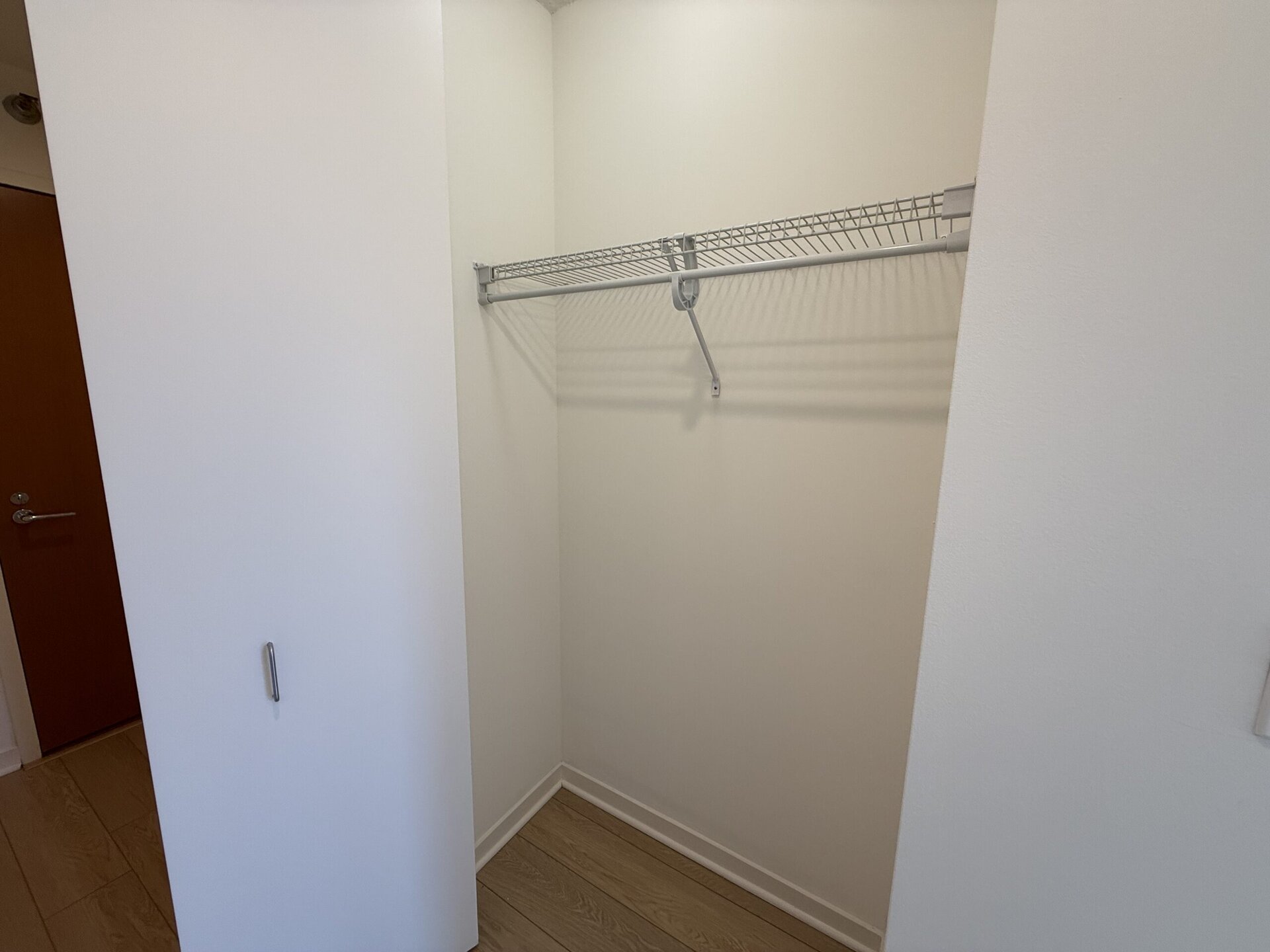 A small, empty closet features a wire shelving unit mounted on the back wall. The shelving is designed for hanging clothes, with a single horizontal rod and a support bracket. The walls are painted white, and the floor is covered with light-colored wood laminate. A closed door is visible on the left side of the image, adding to the minimalistic appearance of the space.