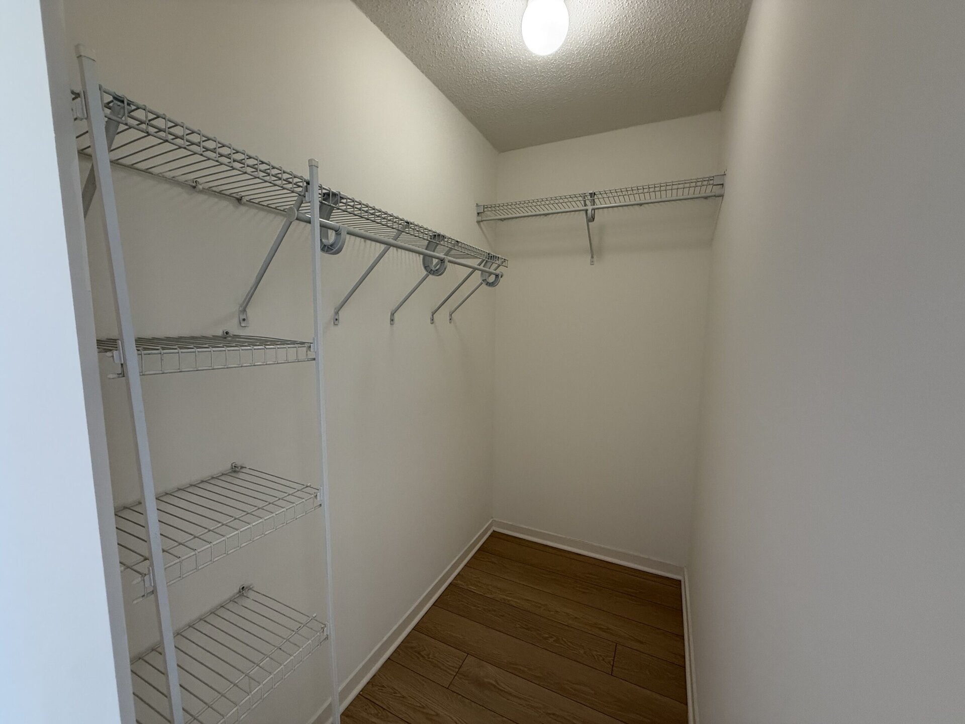 A small, empty closet features white walls and a wooden floor. It has multiple wire shelving units mounted on the walls, with one shelf positioned high and several lower shelves. There are also hooks for hanging items along one wall, creating a functional storage space.