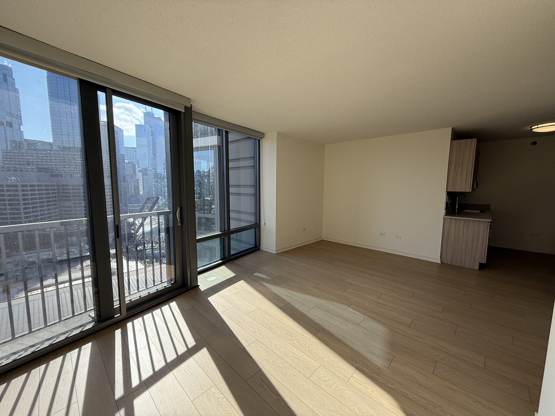 A spacious, modern living area features large glass sliding doors that open to a balcony, offering a view of surrounding skyscrapers. The floor is made of light-colored wood, and the walls are painted in a neutral tone. To the right, there is a small kitchenette with wooden cabinetry and a countertop. Natural light fills the space, creating a bright and airy atmosphere.