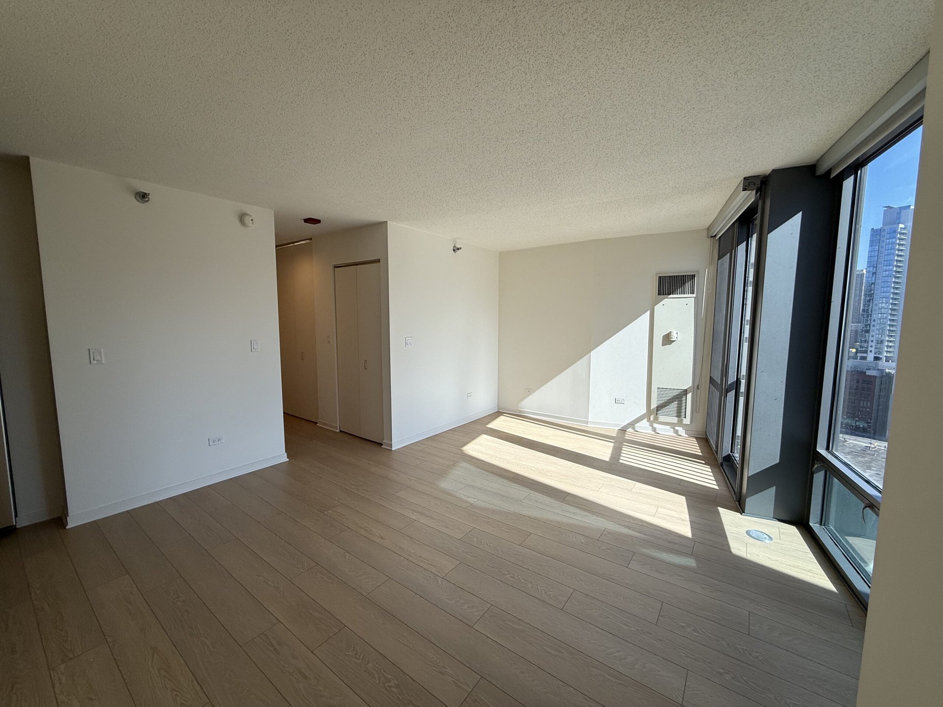 A bright, empty room features light-colored wooden flooring and white walls. Large windows on one side allow natural light to flood in, casting shadows on the floor. There are two closets with sliding doors against one wall, and a small wall-mounted unit is visible. The overall atmosphere is open and airy, with a modern feel.