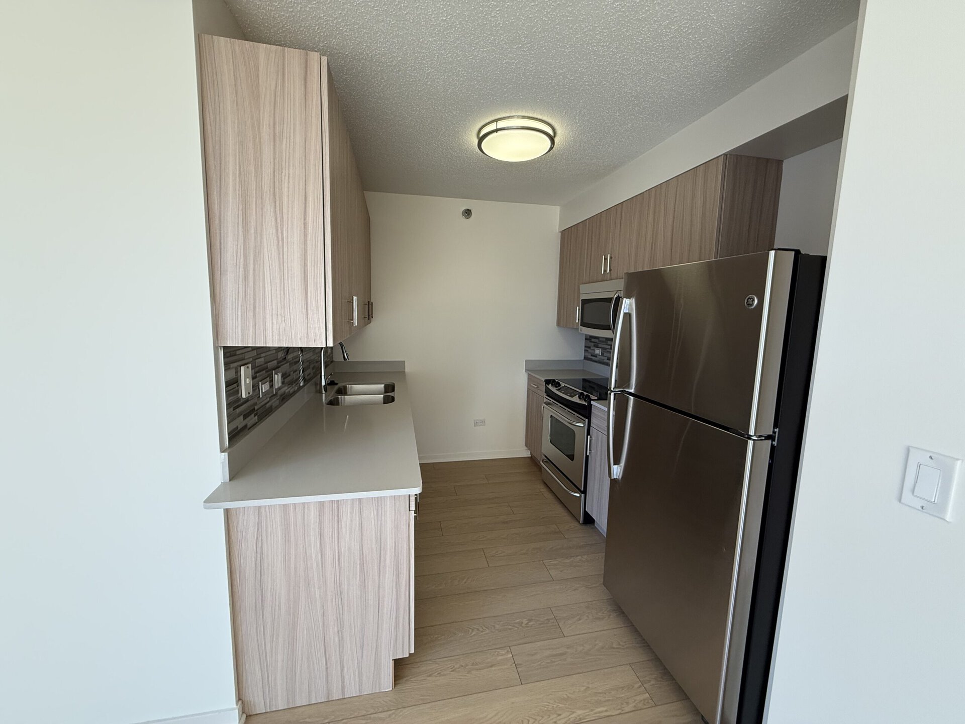 A modern kitchen features light wood cabinetry and a sleek countertop. The space includes stainless steel appliances: a refrigerator, an oven, and a microwave. A double sink is positioned under a window, with a decorative backsplash behind it. The flooring is light-colored, enhancing the bright and airy feel of the room. A ceiling light fixture provides illumination.