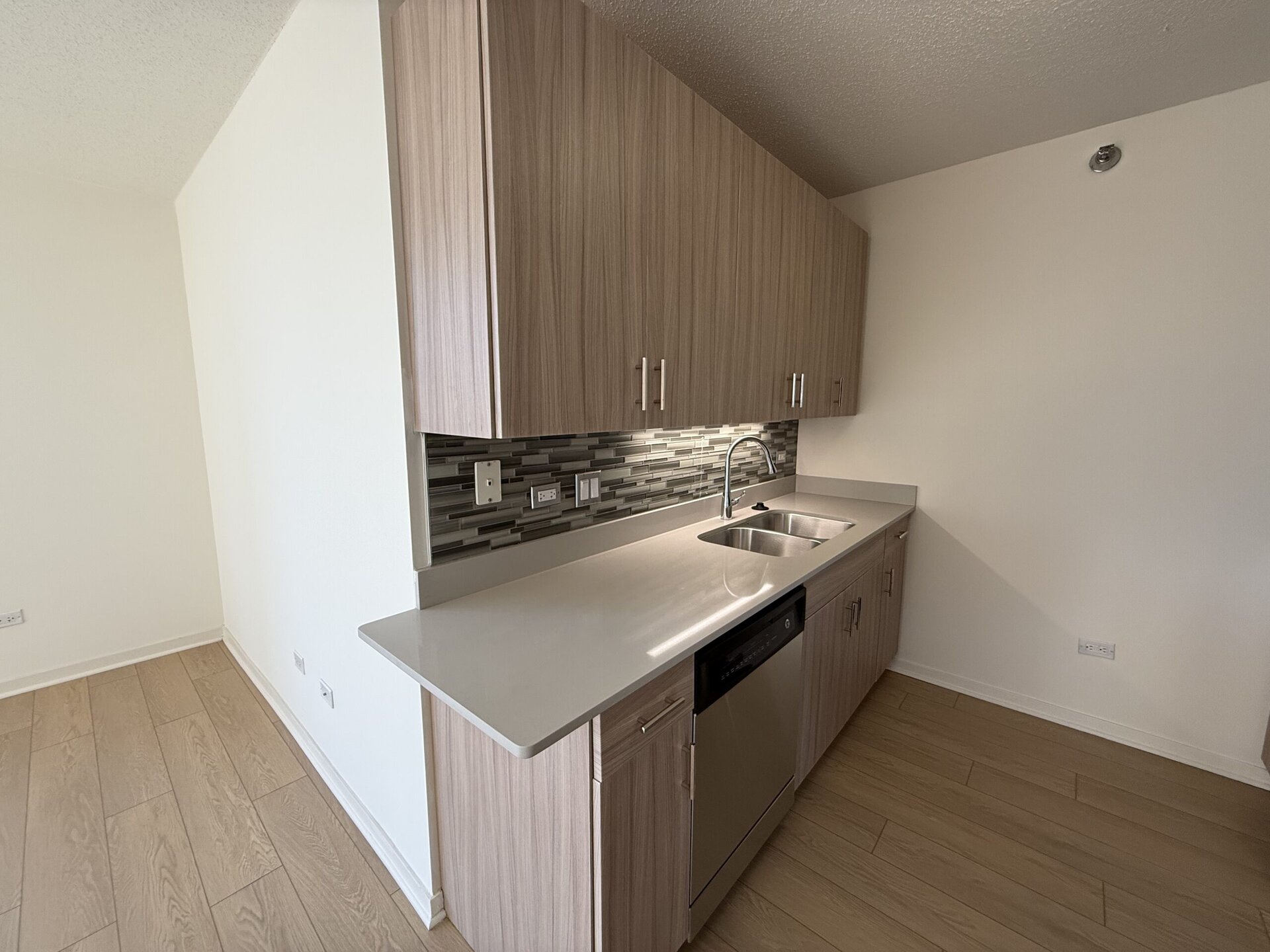 A modern kitchen features light wood cabinetry with sleek handles, set against a stylish backsplash of gray and black tiles. A stainless steel double sink is positioned on a smooth, light-colored countertop, alongside a built-in dishwasher. The flooring is a light wood finish, and the walls are painted white, creating a bright and airy atmosphere.