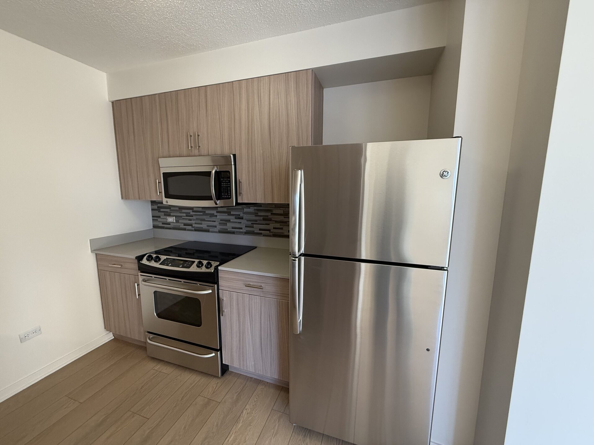 A modern kitchen featuring light wood cabinetry and stainless steel appliances. The setup includes a microwave above a gas stove with an oven, and a refrigerator to the right. The backsplash is composed of gray and white tiles, complementing the light countertops. The flooring is a light wood finish, contributing to a bright and contemporary aesthetic.