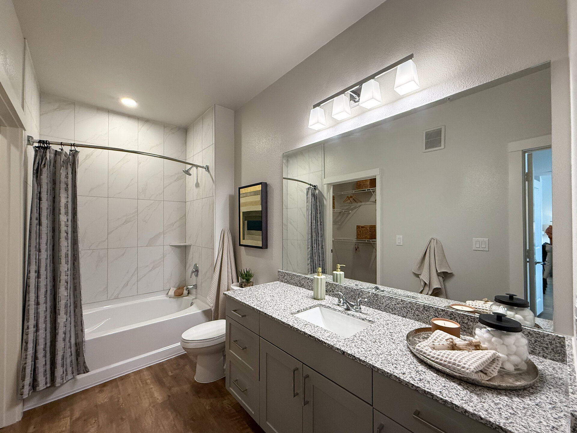 A modern bathroom featuring a garden-style bathtub with a shower curtain, set against a wall of light-colored tiles with subtle veining. The countertop is made of speckled granite, with a rectangular sink and a sleek faucet. Above the sink, a large mirror reflects the space, illuminated by a row of contemporary light fixtures. A door leads to a closet area with shelves and storage baskets.